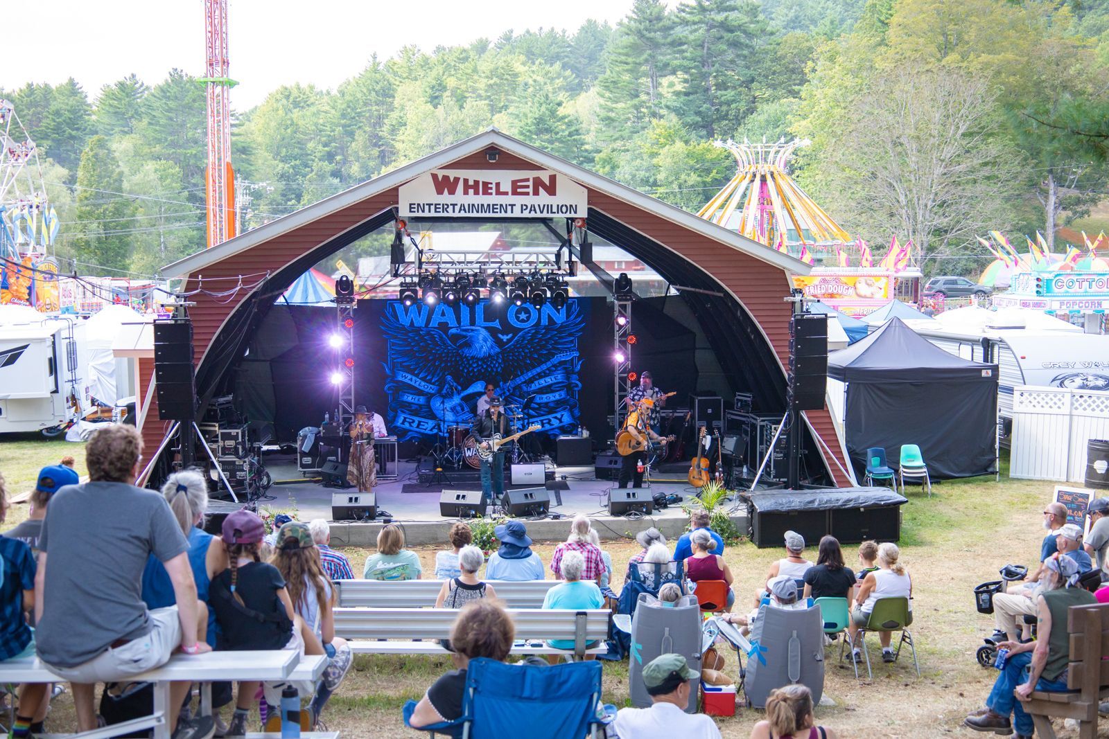 Outdoor music festival with a lit stage, seated audience, and colorful rides in the background