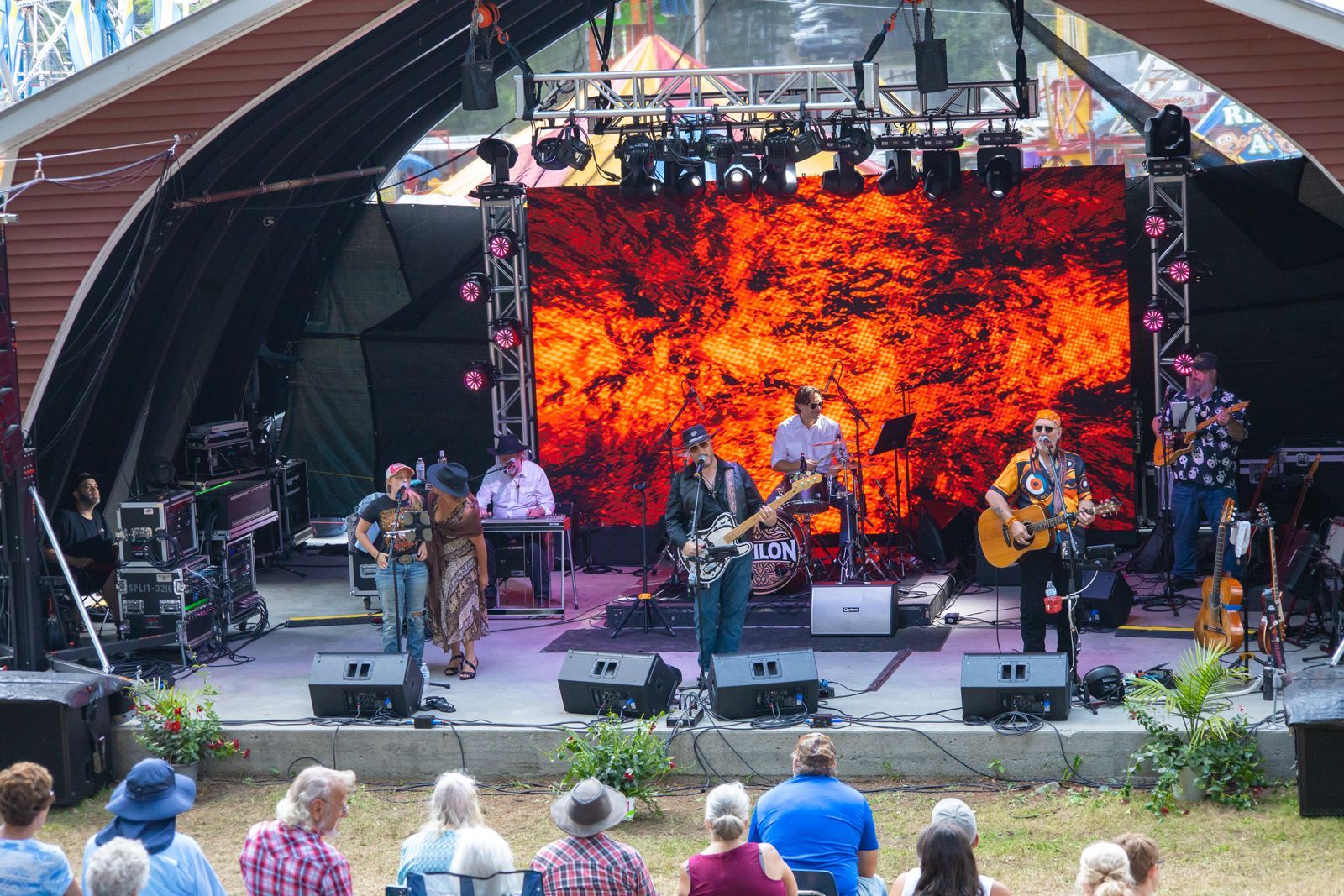 Outdoor band performing on a stage with a bright orange-red screen behind them, audience seated in front