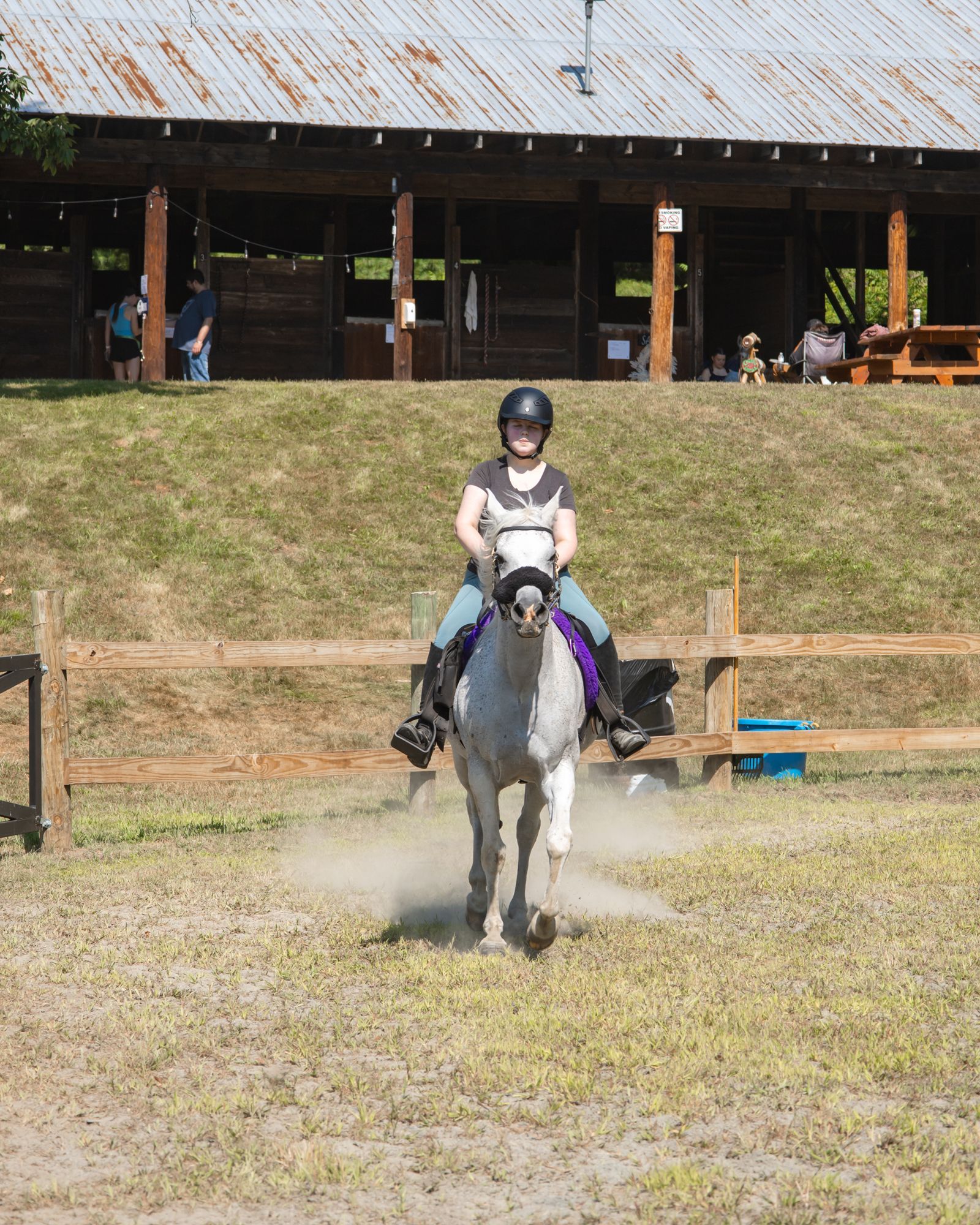 Rider on a gray horse trotting in an outdoor arena, with a barn and spectators in the background