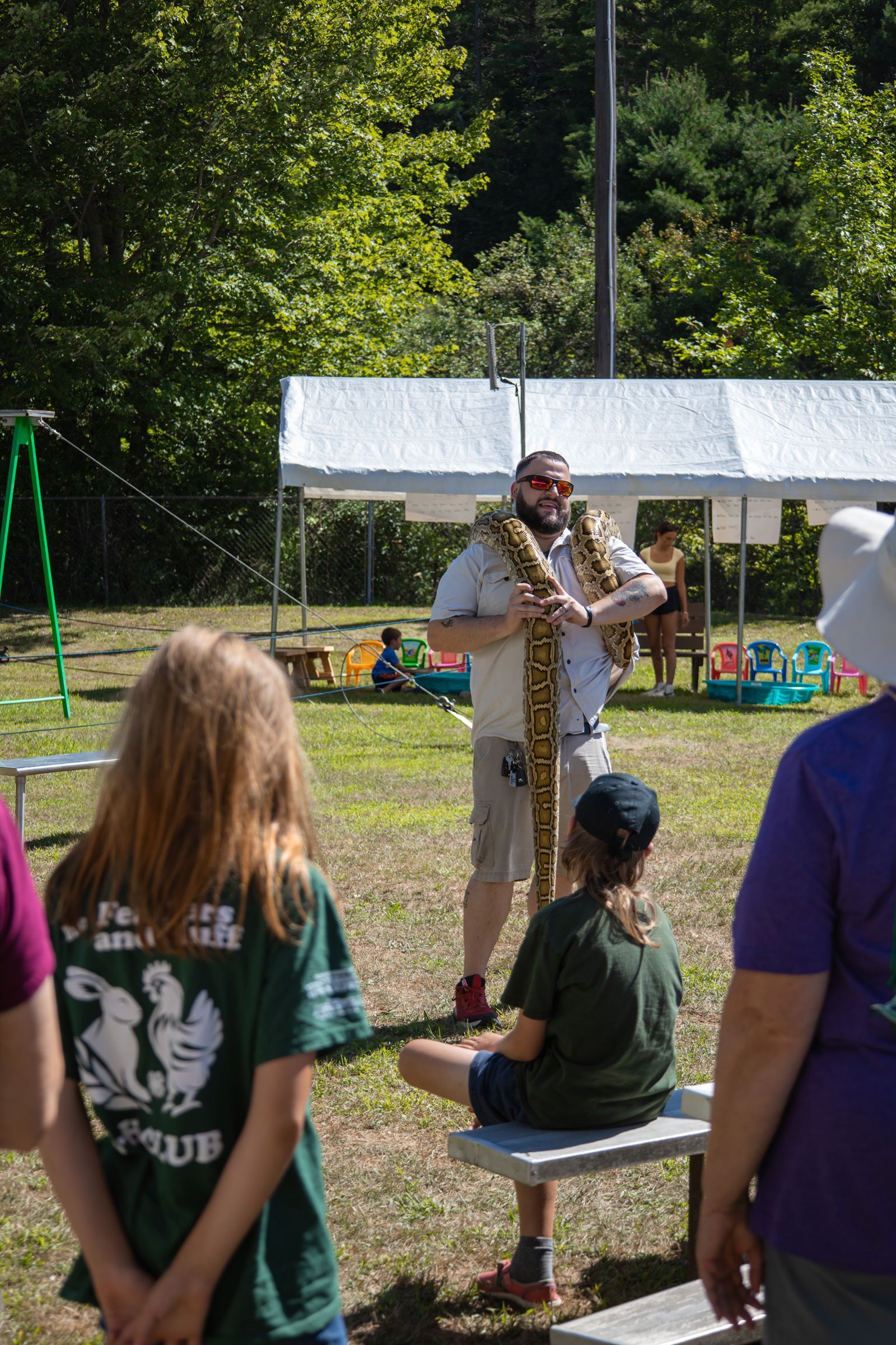 Person speaking into a microphone at an outdoor event, with a white tent and children watching nearby