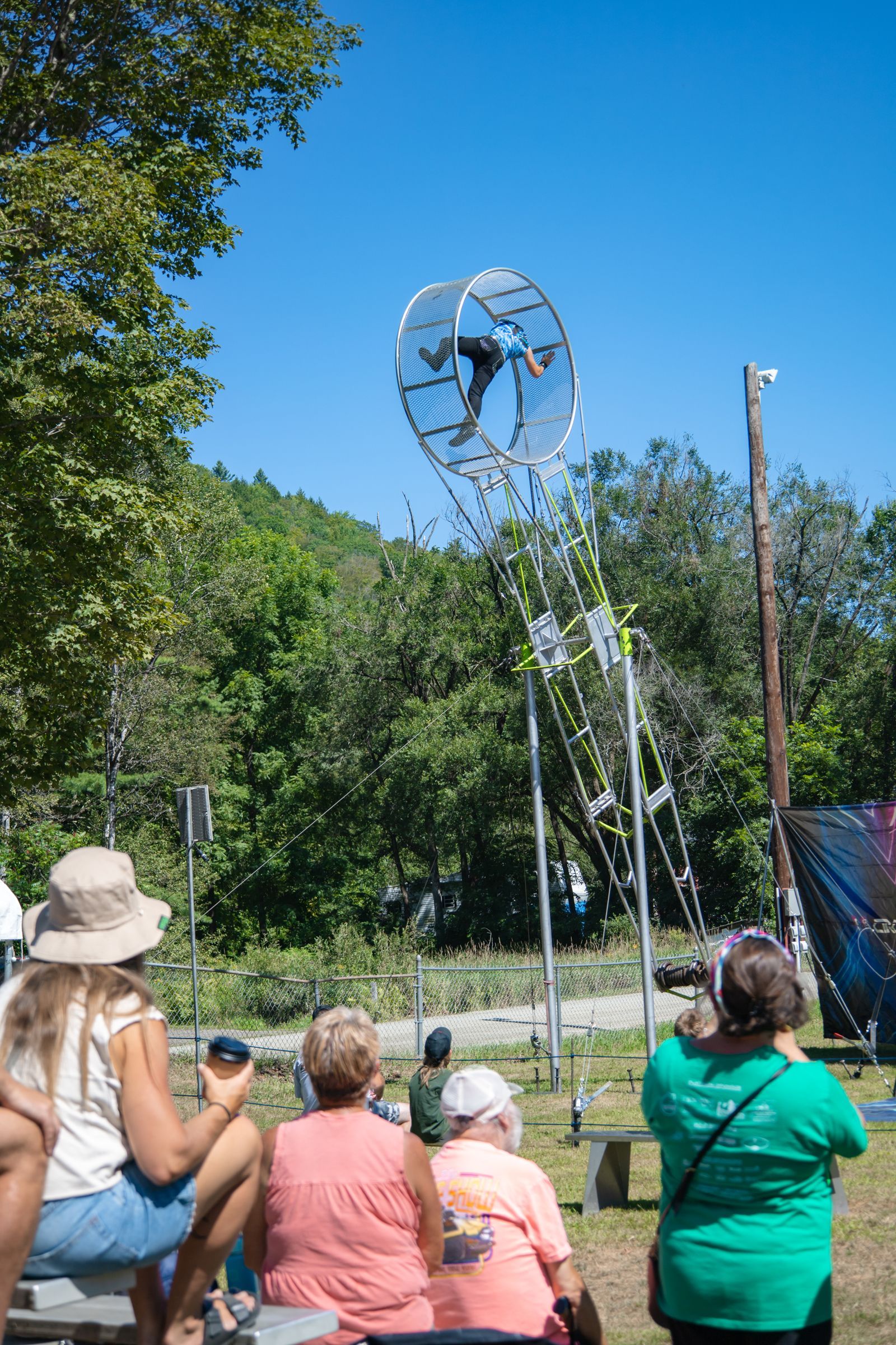 People sitting outdoors watch a tall metal kinetic sculpture against a blue sky.