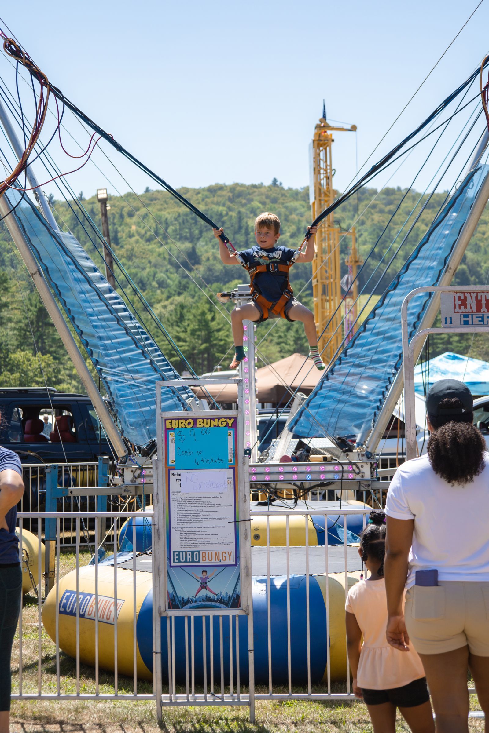 Child on blue bungee trampoline at an outdoor fair, with an adult nearby and mountains in the background