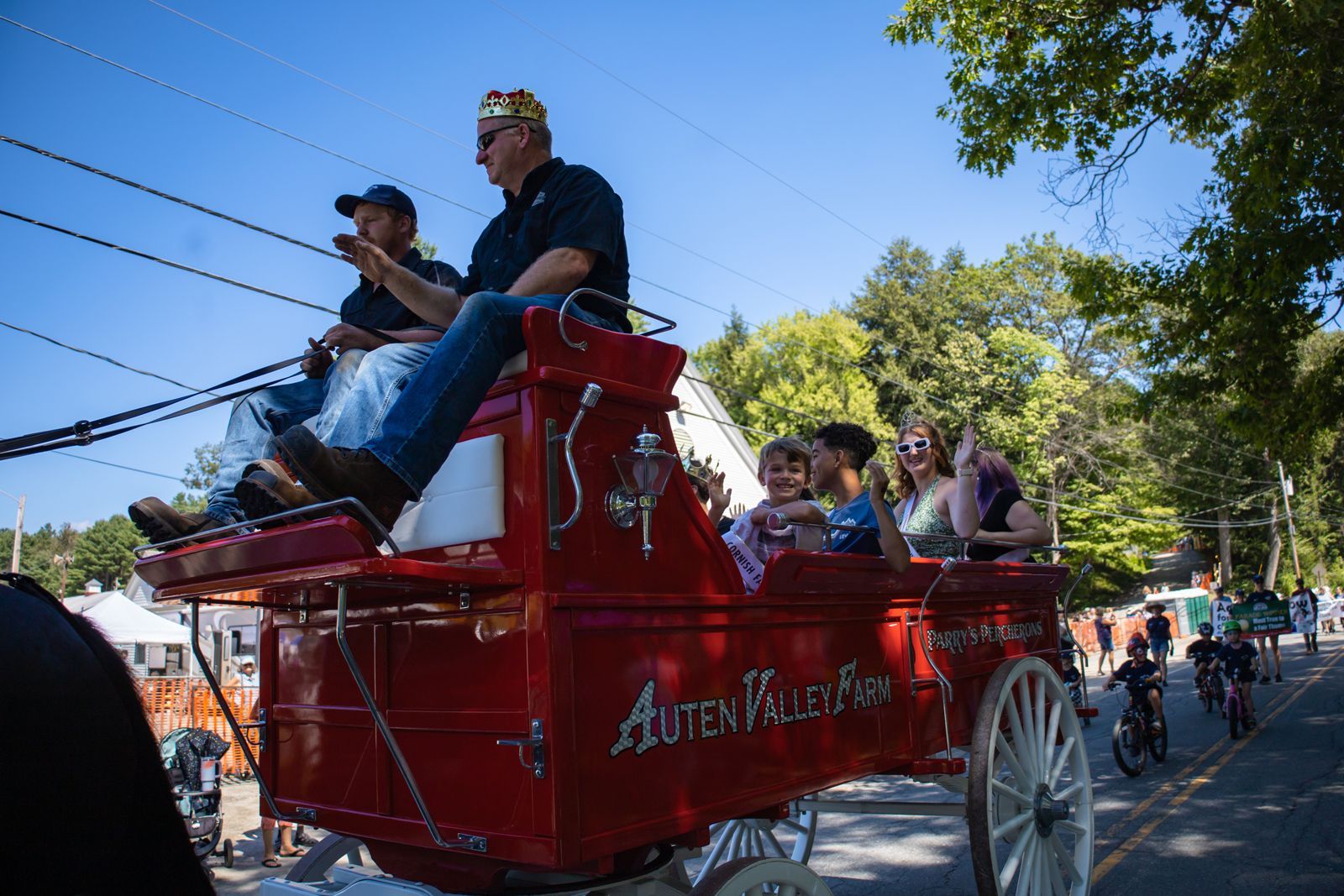 People riding in a red horse-drawn parade carriage on a sunny street, with a crowd in the background