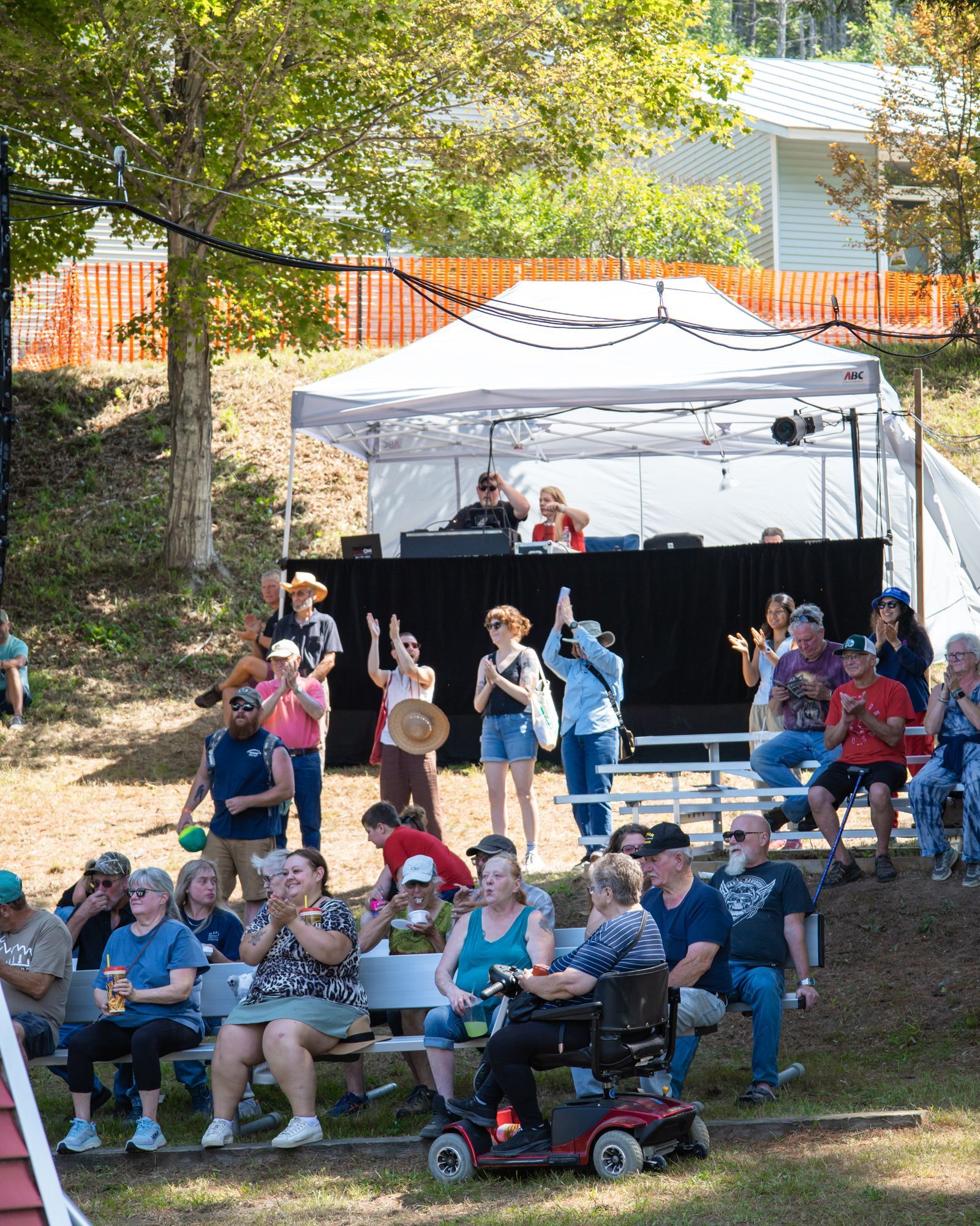 Outdoor crowd seated on a grassy slope at a park event under a white canopy stage
