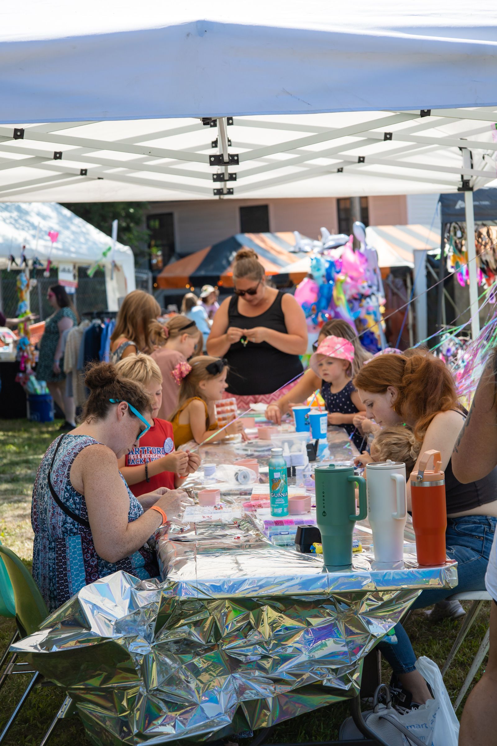People crafting at a shiny table under a white canopy at an outdoor fair