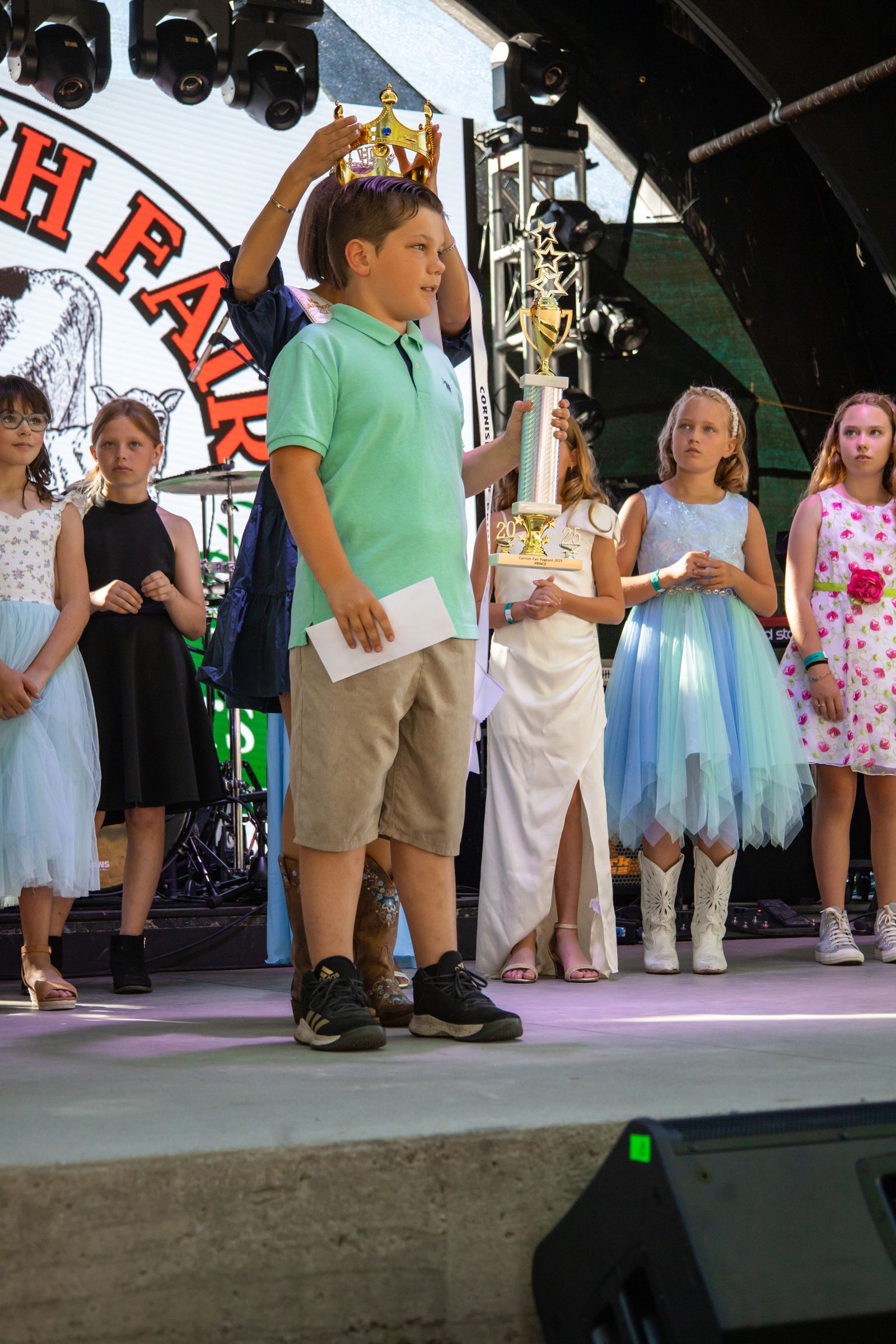 Children standing on a stage at an outdoor event, with one boy holding a paper in front of a trophy display.