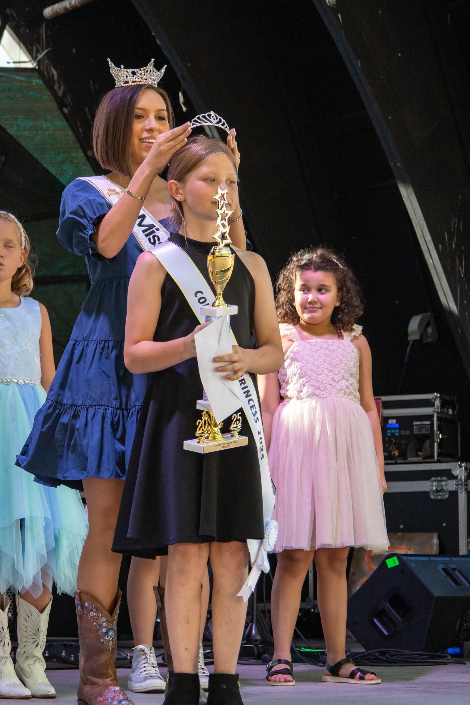 Children on stage, one wearing a crown and sash, with others standing beside her in dresses