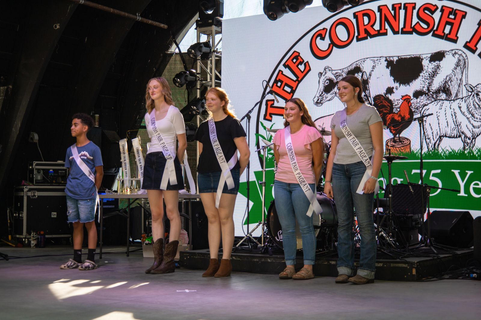 Six performers standing on a stage in front of a Cornish banner, wearing casual and performance outfits.