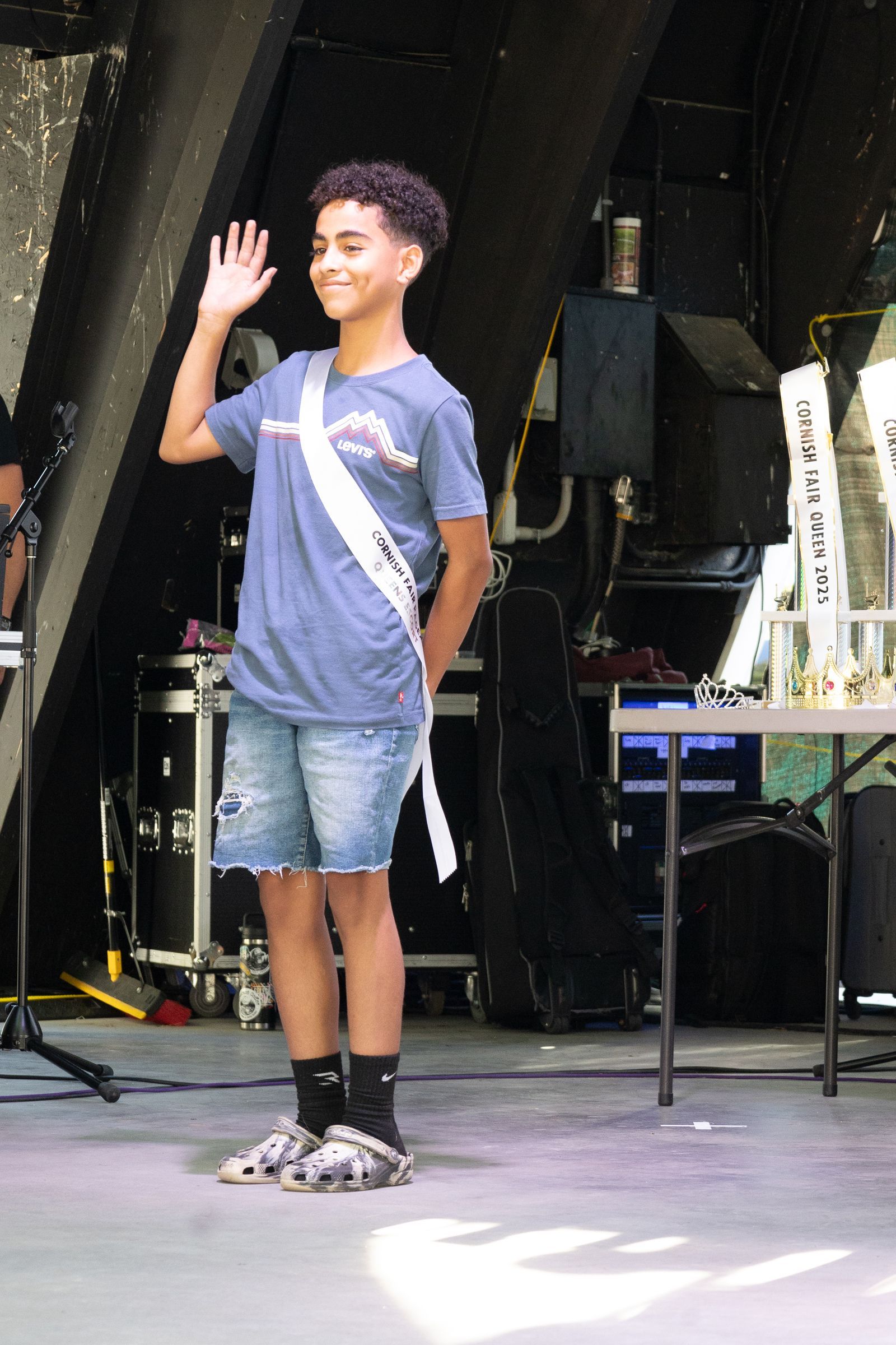 Person in a blue shirt and denim shorts waves on a stage backstage area.