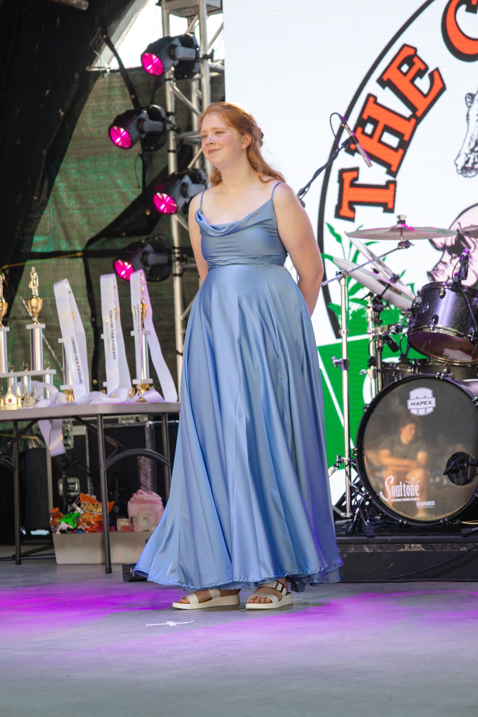 Woman in a blue dress singing on an outdoor stage with drums and purple lights