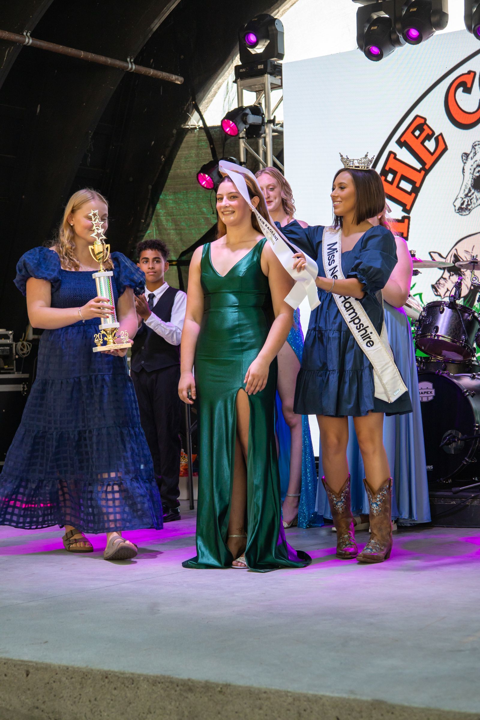 Three women in formal dresses stand onstage under purple lights, one holding a trophy and one speaking at a mic.