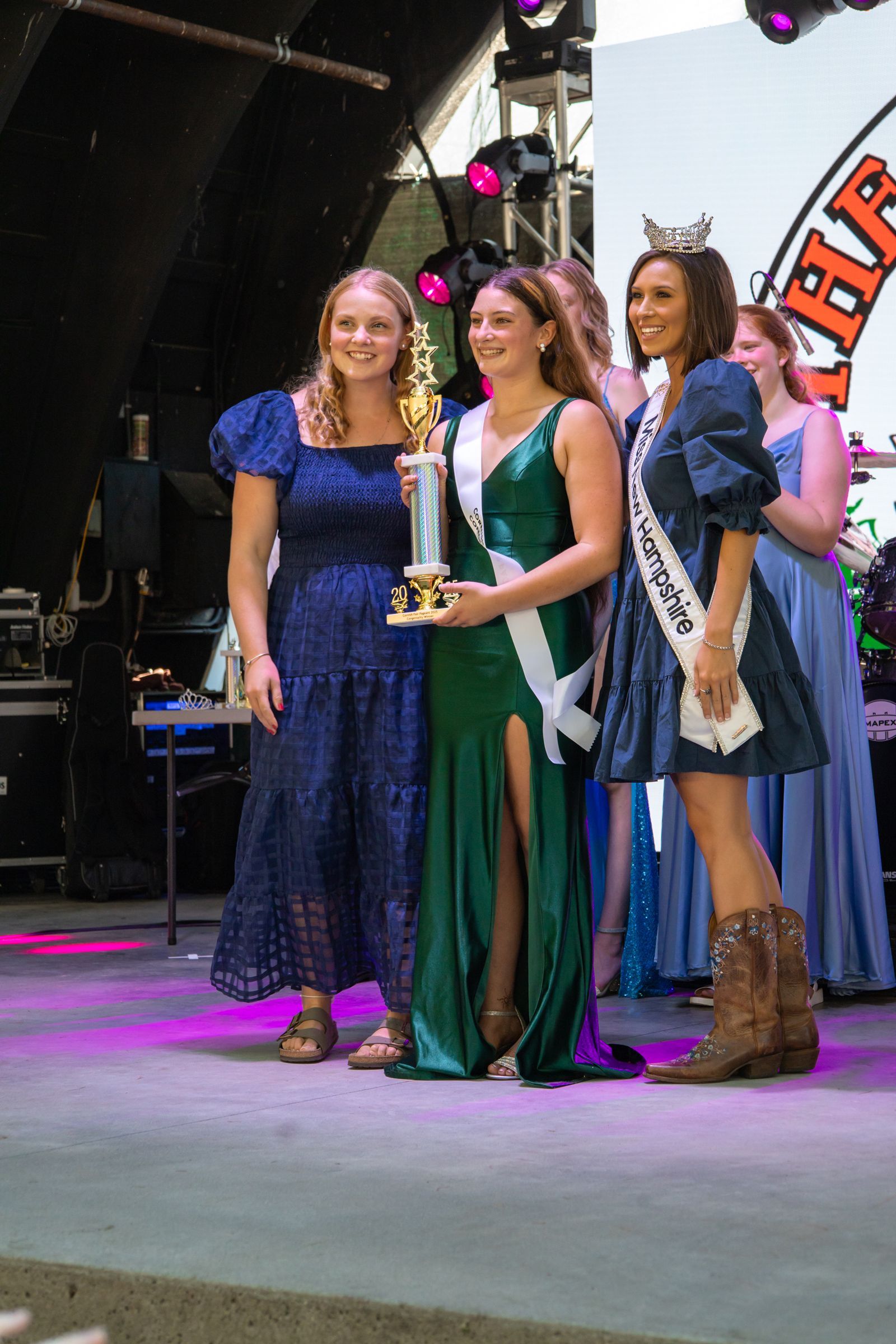 Three women on stage holding a trophy, dressed in formal blue and green gowns under stage lights