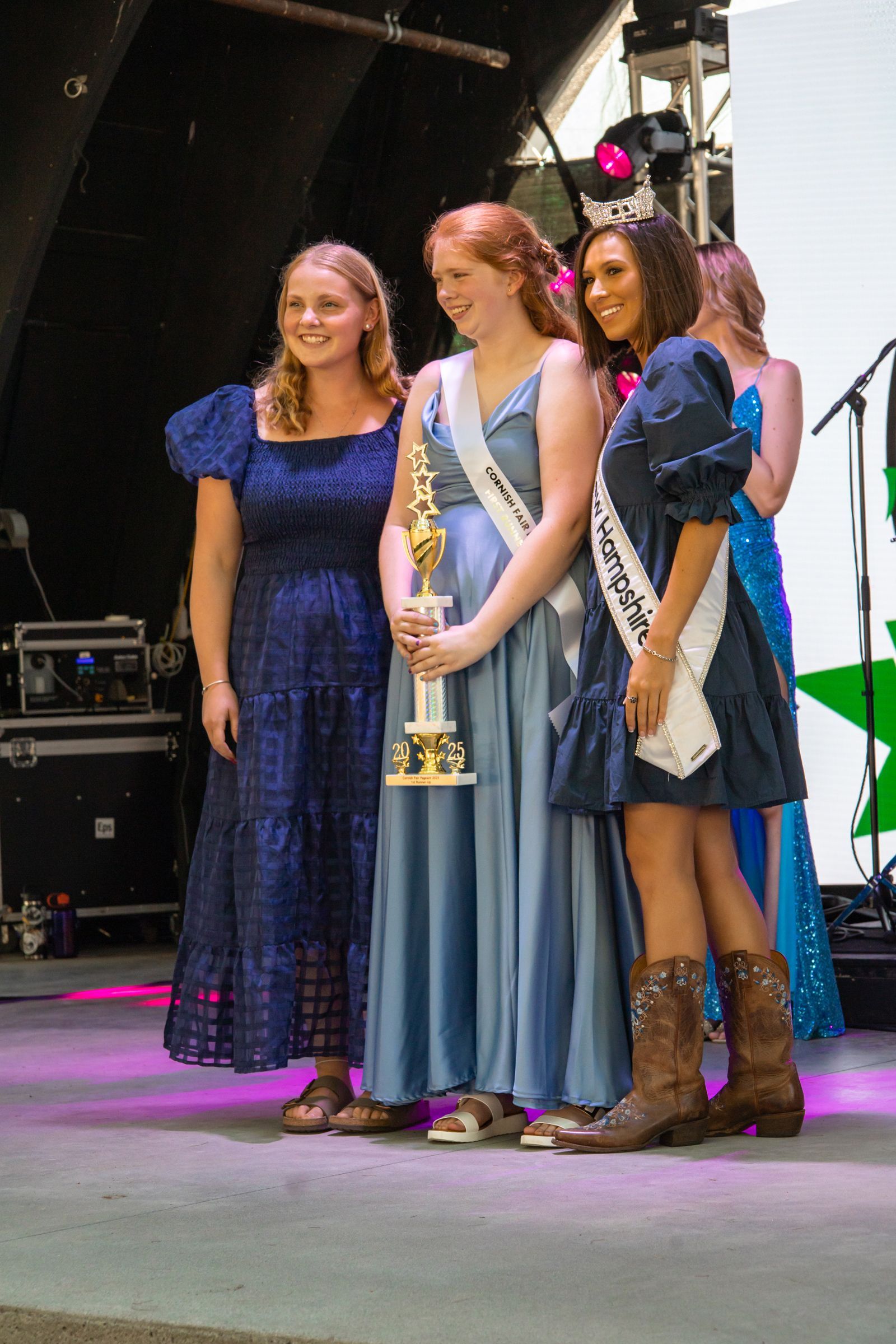 Three women in sashes pose onstage, one holding a trophy, in formal gowns and boots.