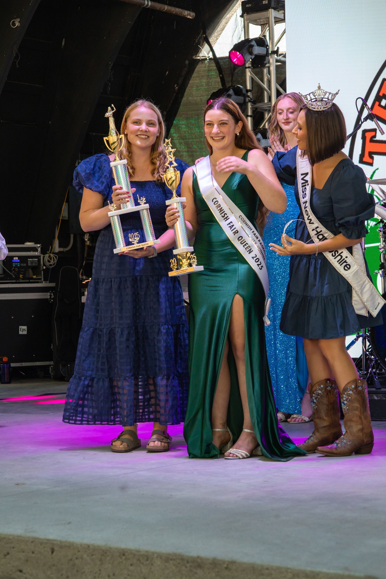 Three women on stage in dresses, one holding a trophy and two wearing sashes.