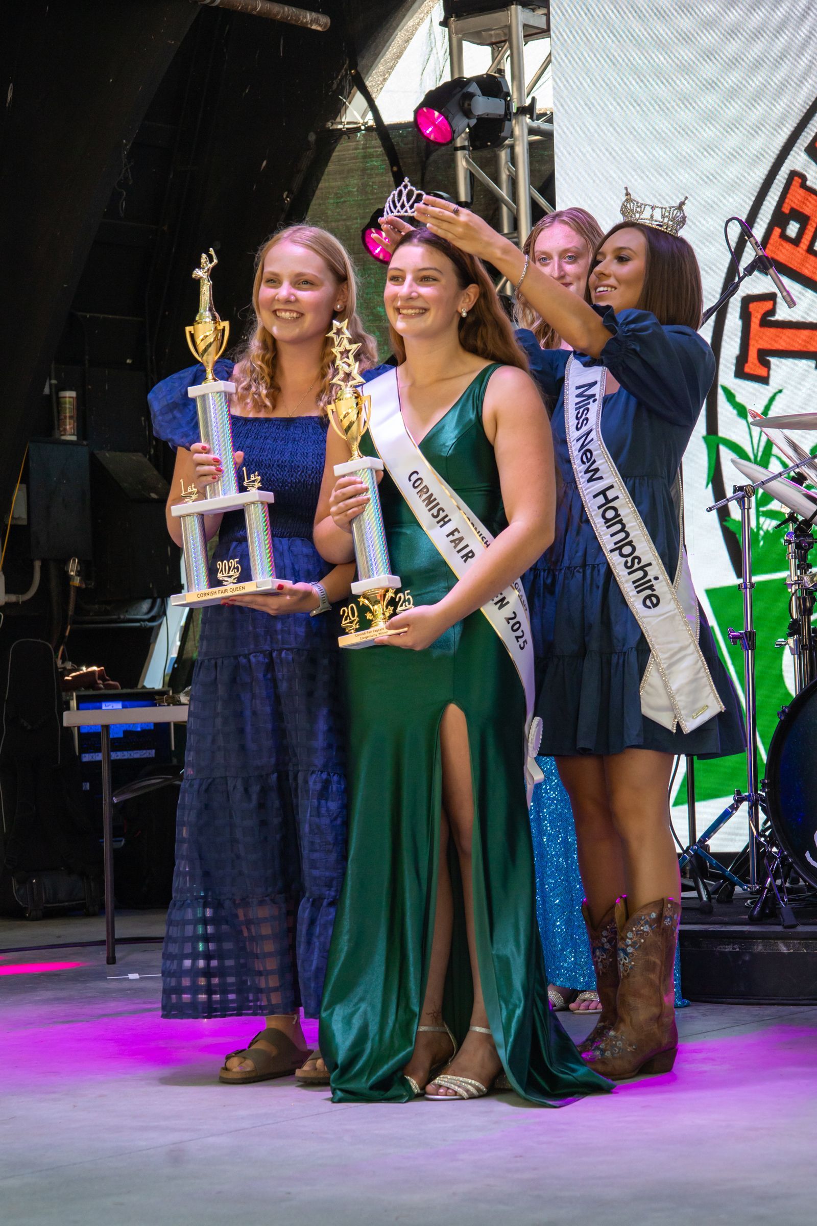 Three women on stage posing with trophies, one in a green gown and two in blue dresses, under stage lights