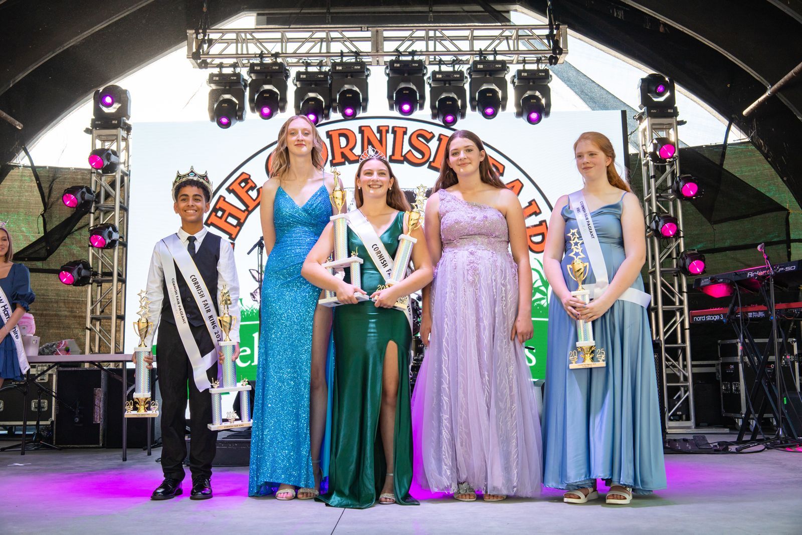 Six people in colorful gowns on an outdoor stage, standing under a carnival-style sign.