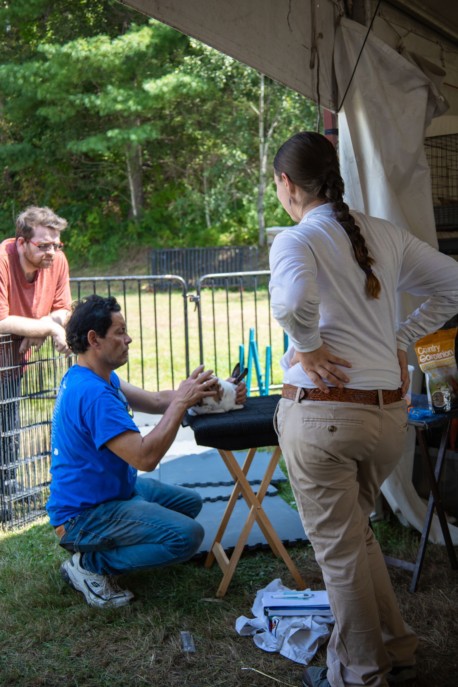 People chatting at a tented outdoor gathering, with one person kneeling by a table and bottles visible.