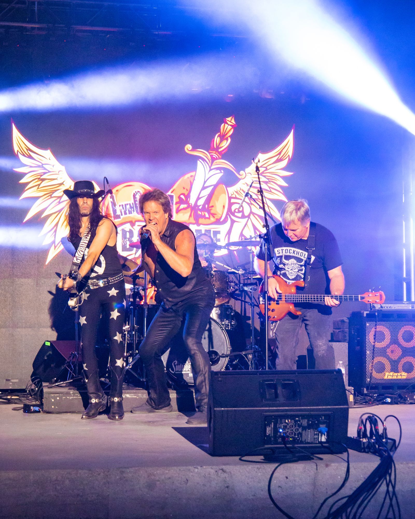 Rock band performing onstage under blue lights, with drums, guitars, and winged backdrop.