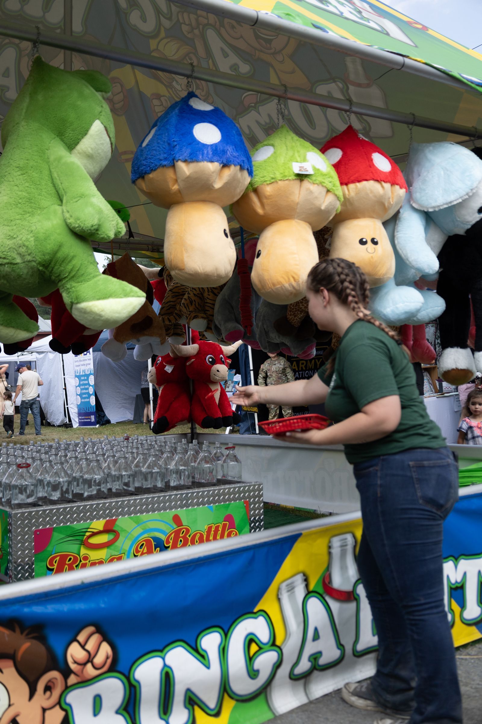 Woman at carnival booth reaching for hanging plush toys, including green, blue, yellow, and red ones.