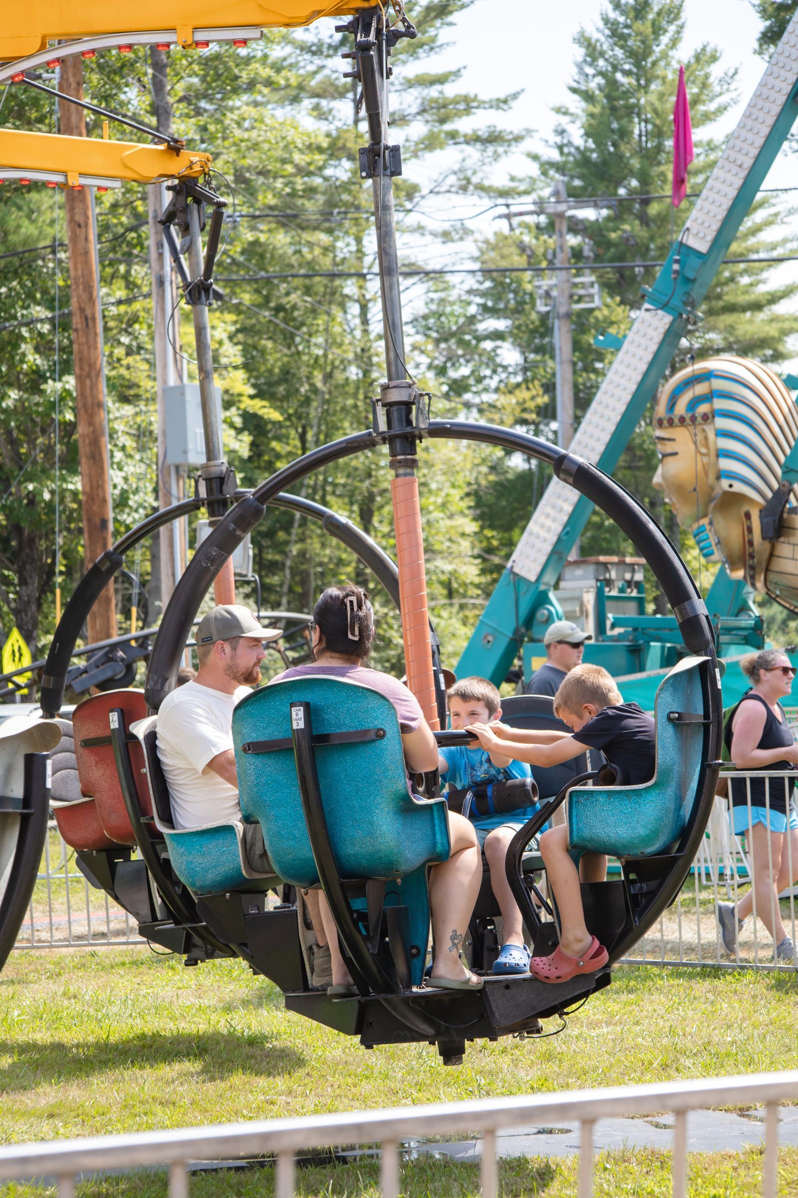 People riding a round amusement ride outdoors, with blue seats and trees in the background
