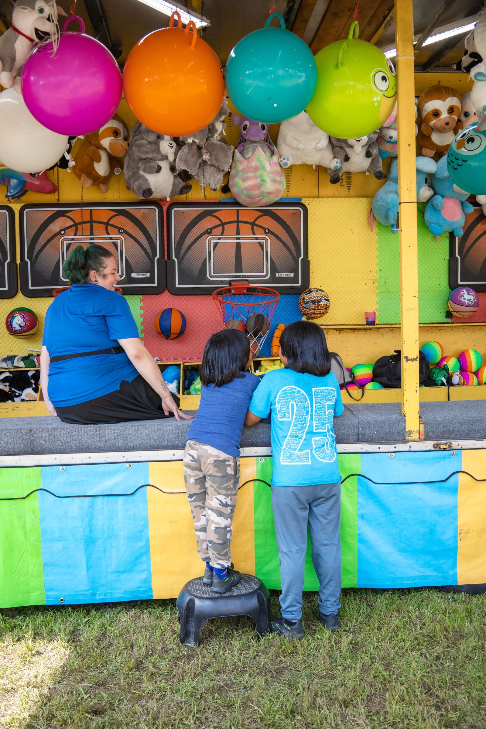 Children at a colorful carnival game booth reaching toward prizes and balloons