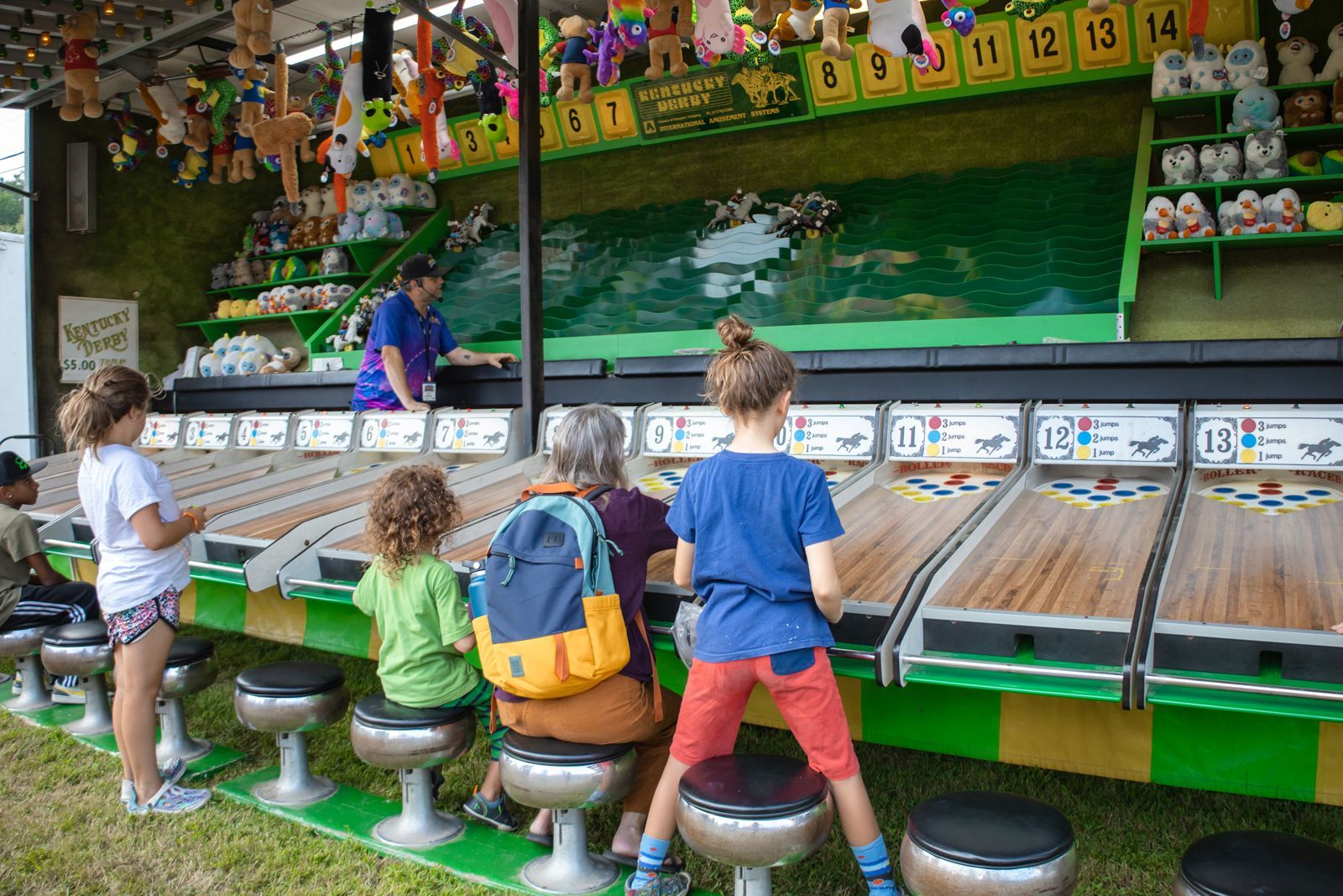 Children sitting on stools at a carnival fishing game booth with colorful prizes and a green backdrop