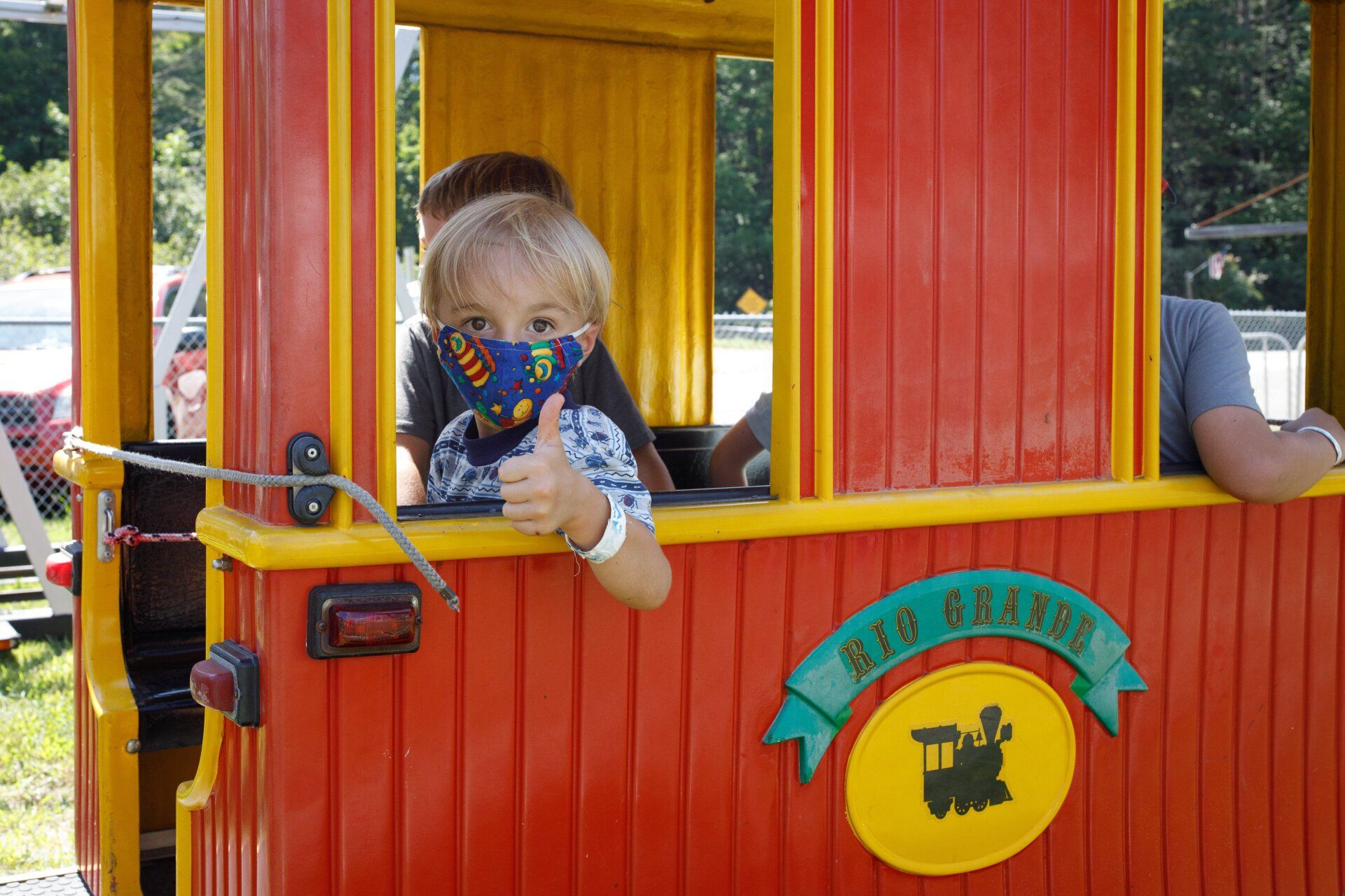 A little boy wearing a mask is riding a train and giving a thumbs up