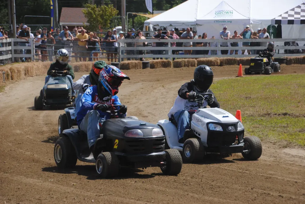 Two riders race side-by-side on ATVs on a dirt track, with spectators behind a fence.