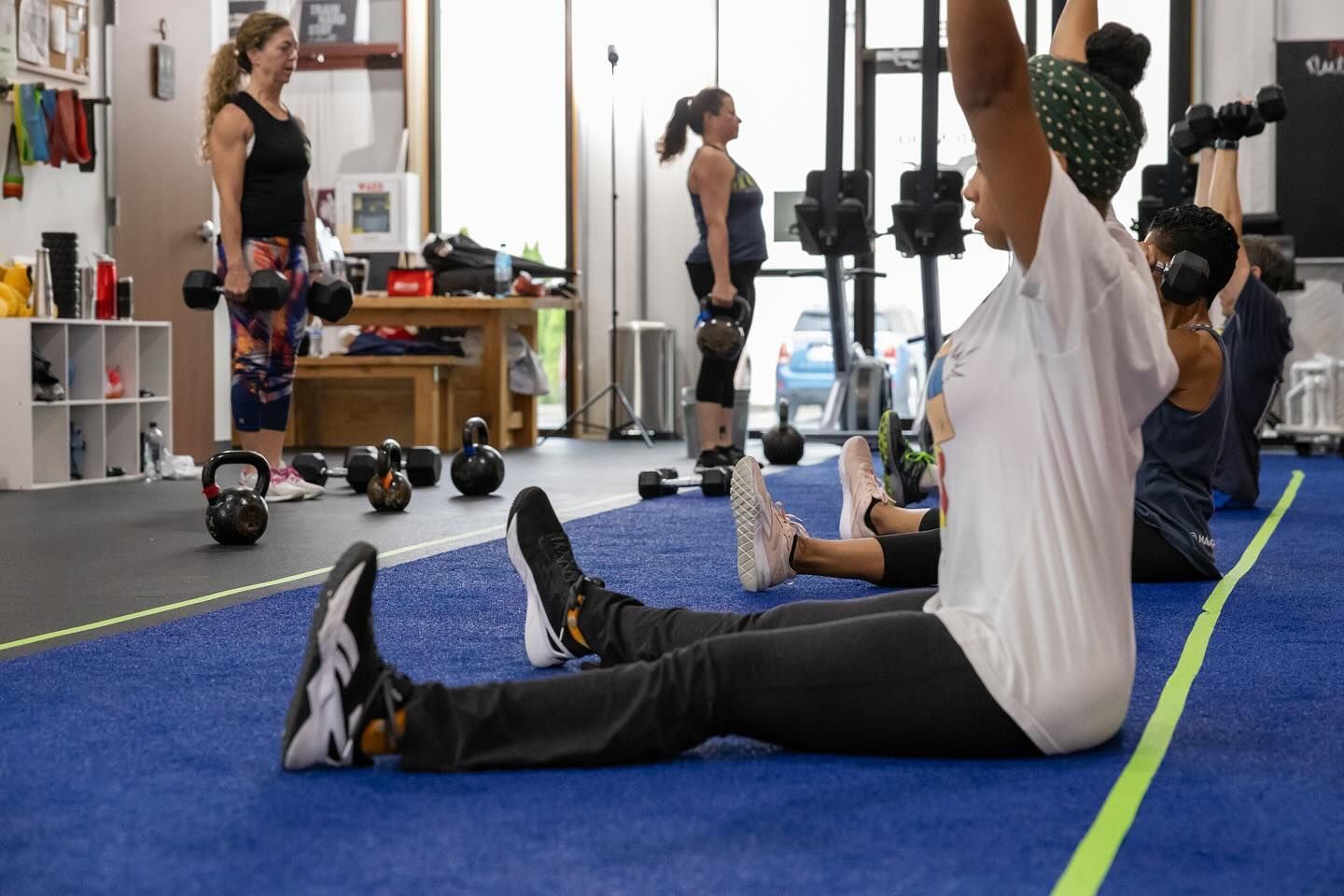 People exercising with weights at Mad Mare CrossFit gym in Alpharetta, GA. Some seated, some standing. A coach observes