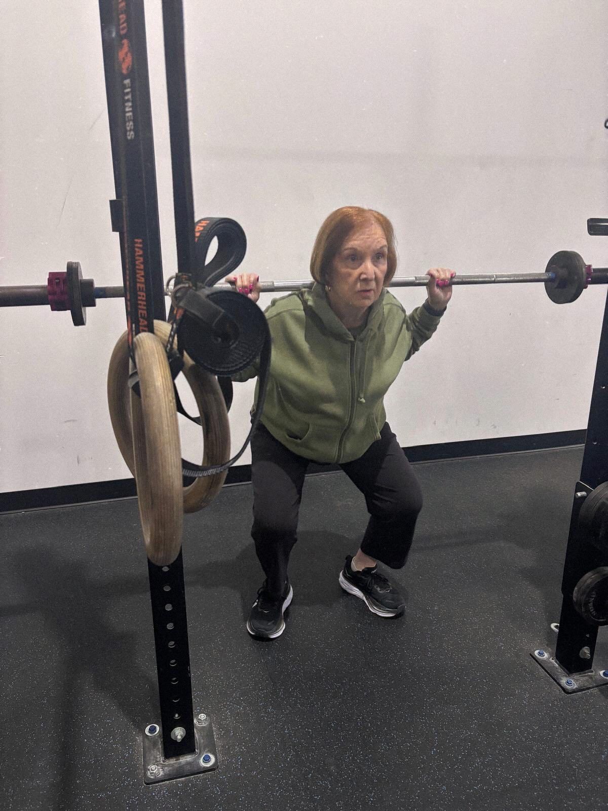 An older person performs a barbell squat at Mad Mare CrossFit gym in Alpharetta, GA in a green hoodie and black pants.