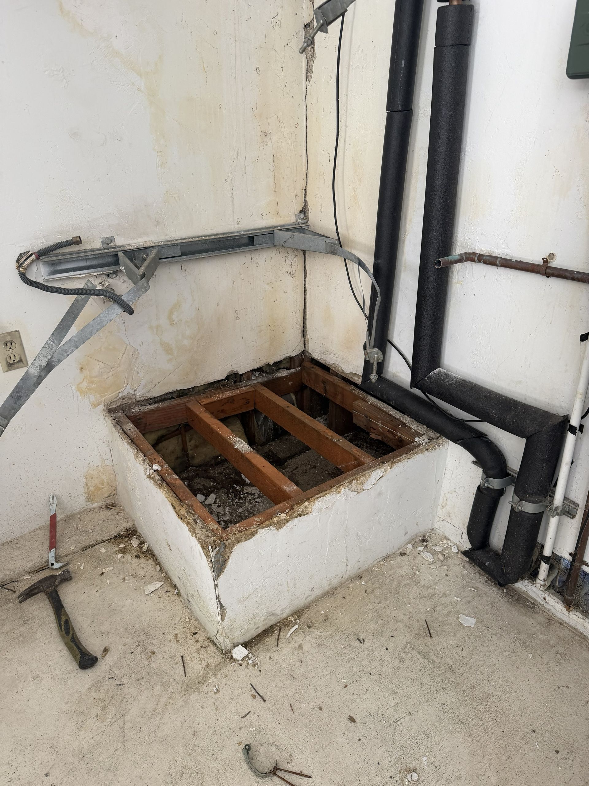 Open floor access hatch with rusted metal frame beside black pipes in a utility room corner