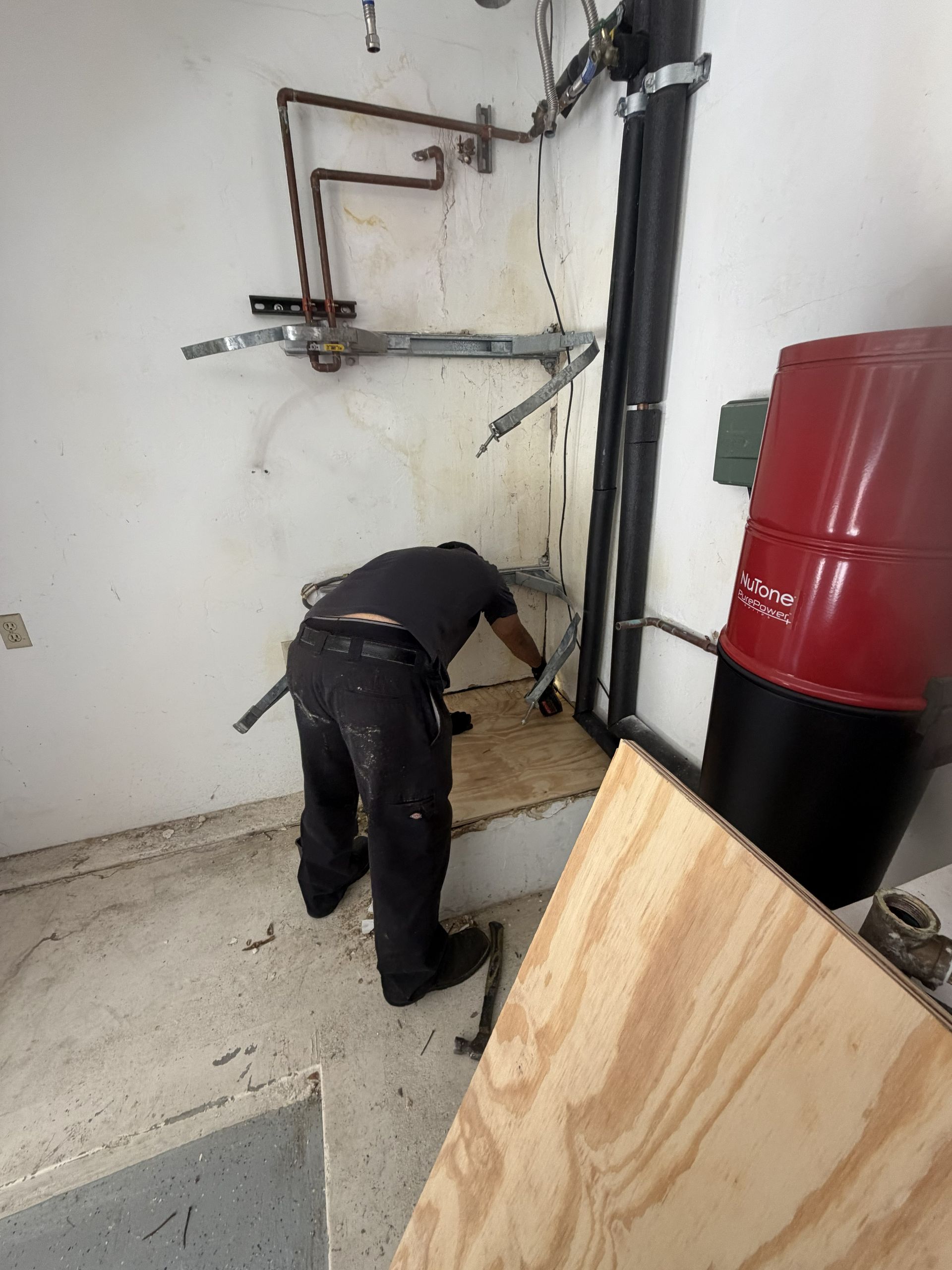 Worker bending near pipes in a basement, beside a red tank and black utility pipe.