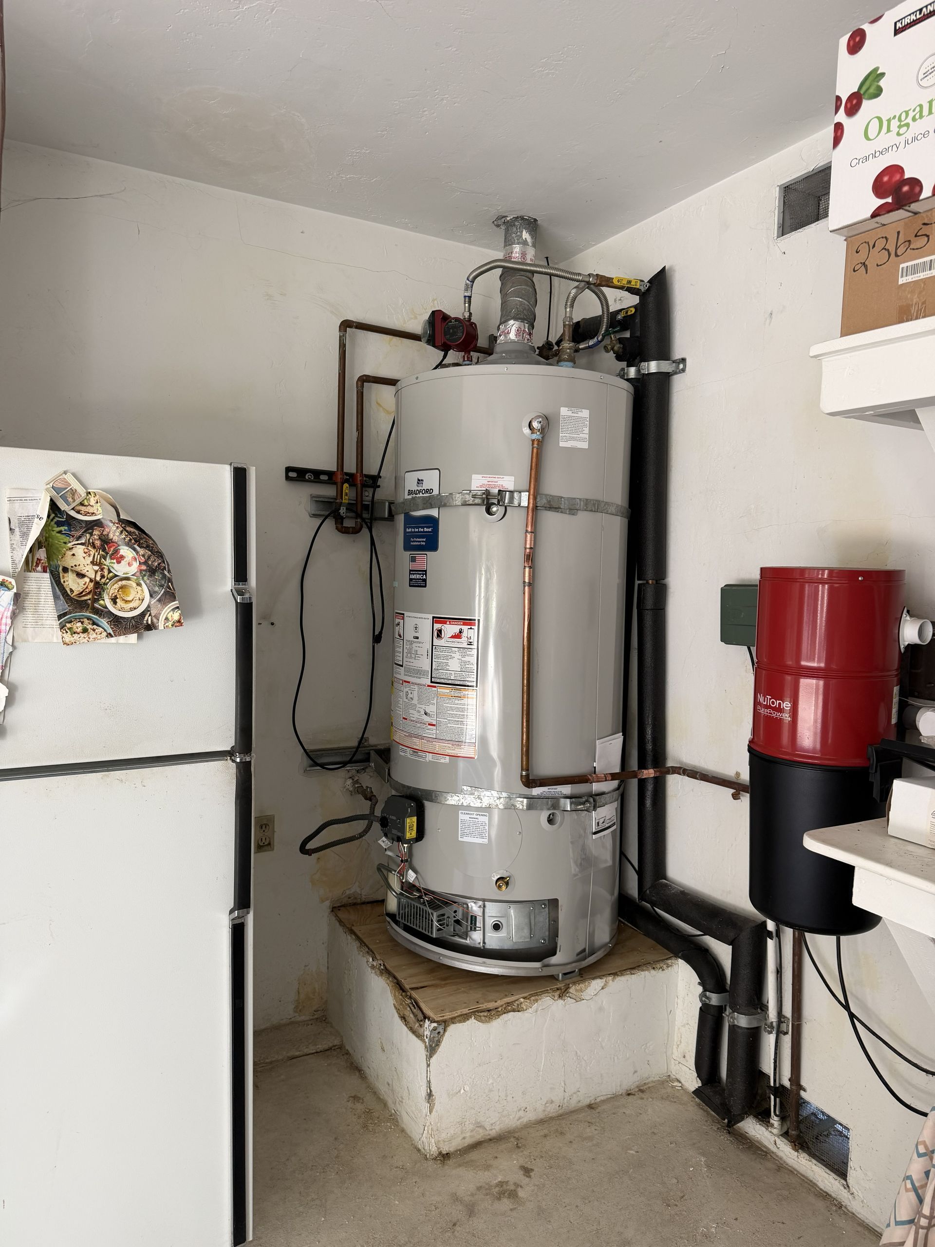 Basement water heater beside a white refrigerator, with red tank and pipes in a utility room