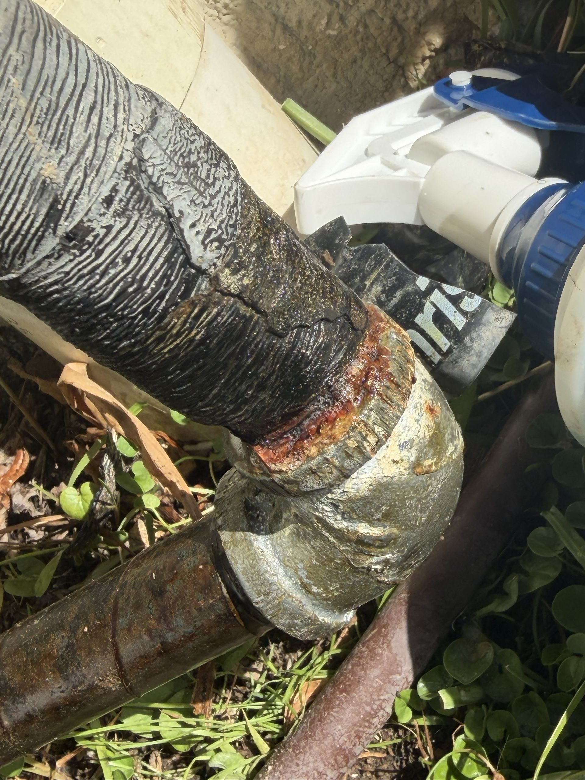 Close-up of a damaged insulated pipe wrapped with tape, outdoors near grass and a white fitting.