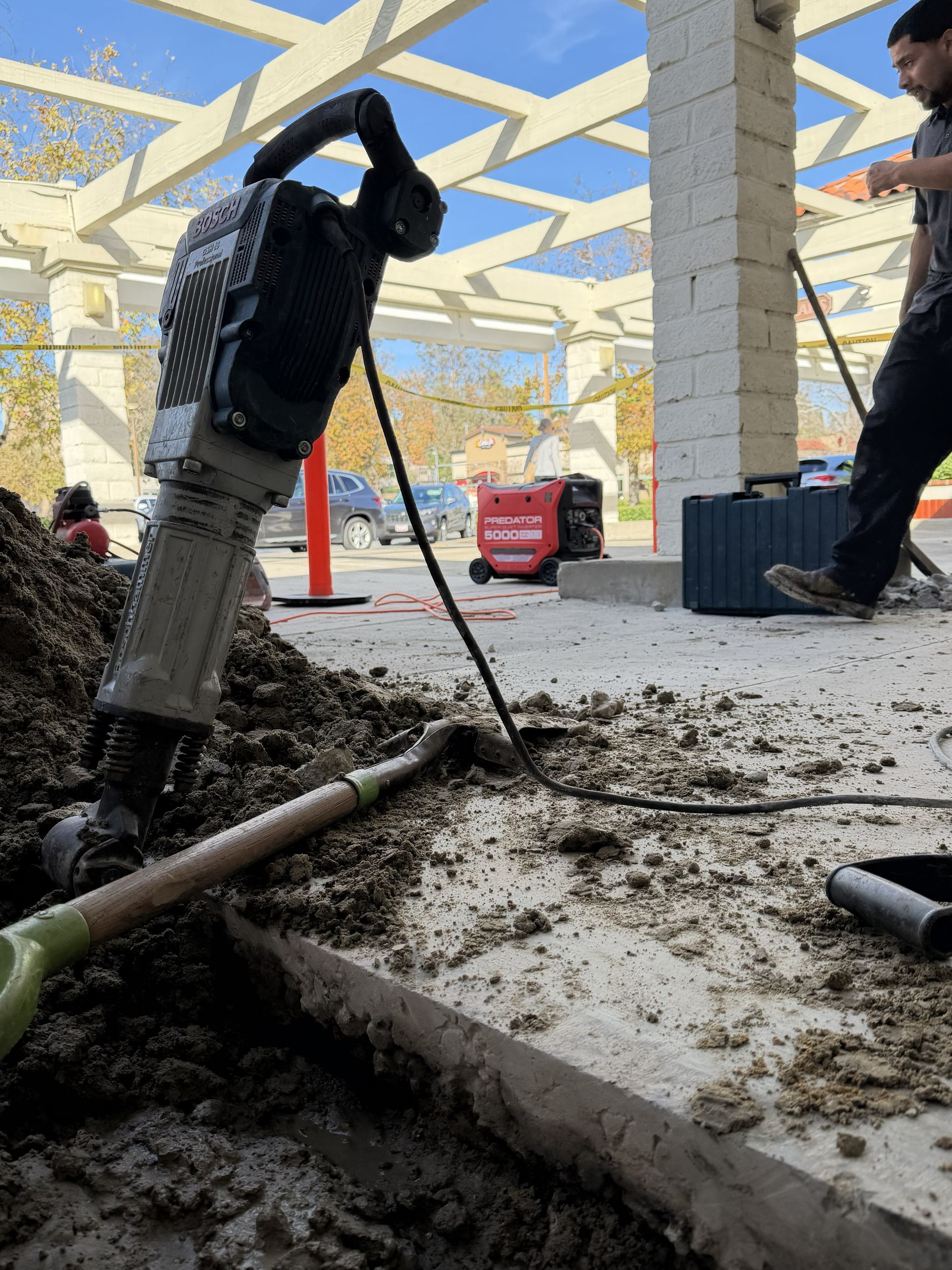Worker using a jackhammer breaks concrete in a construction area under a canopy.