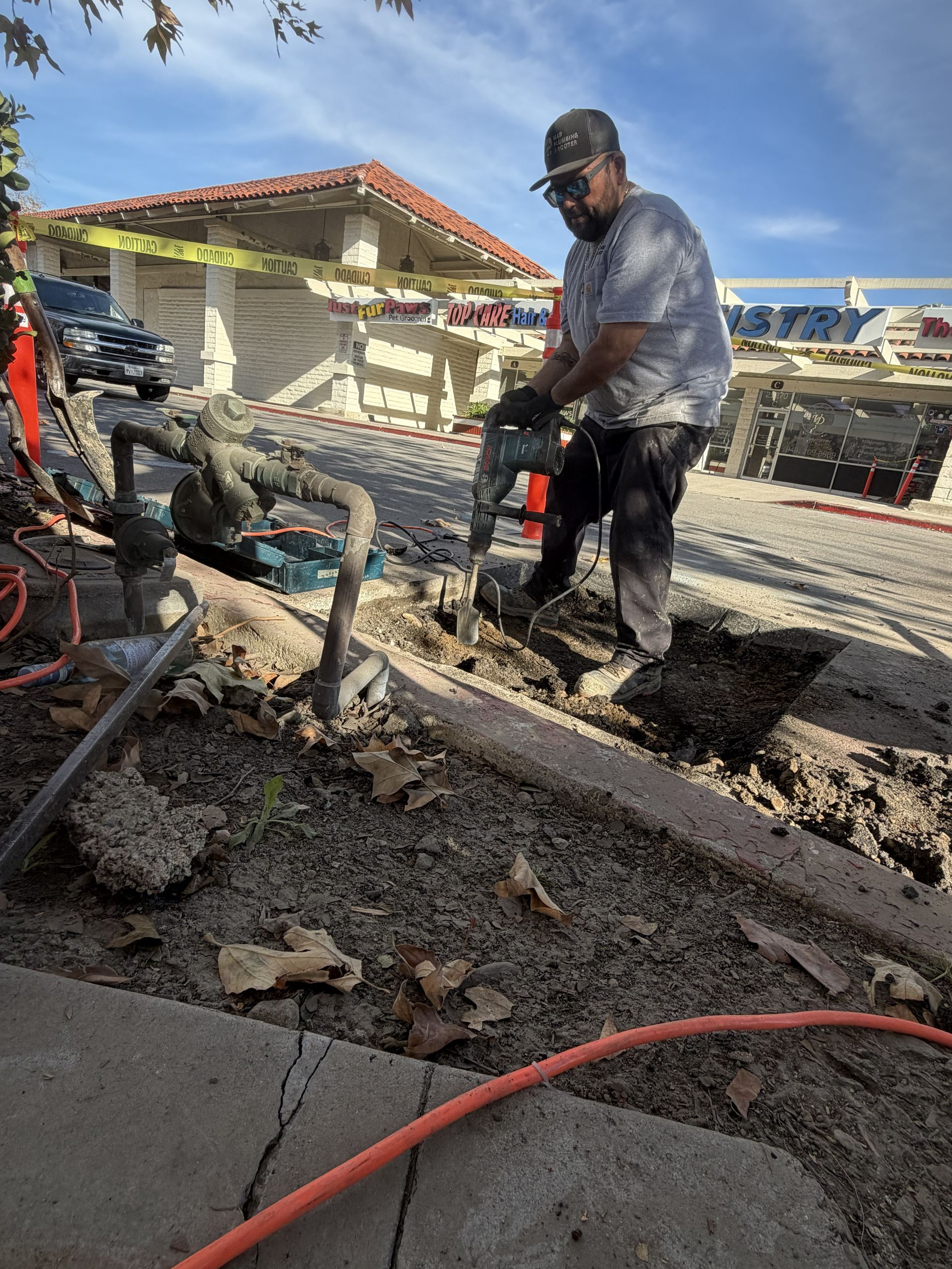 Worker pressure-washes muddy pavement beside a building, with hoses and a red hose in the foreground.