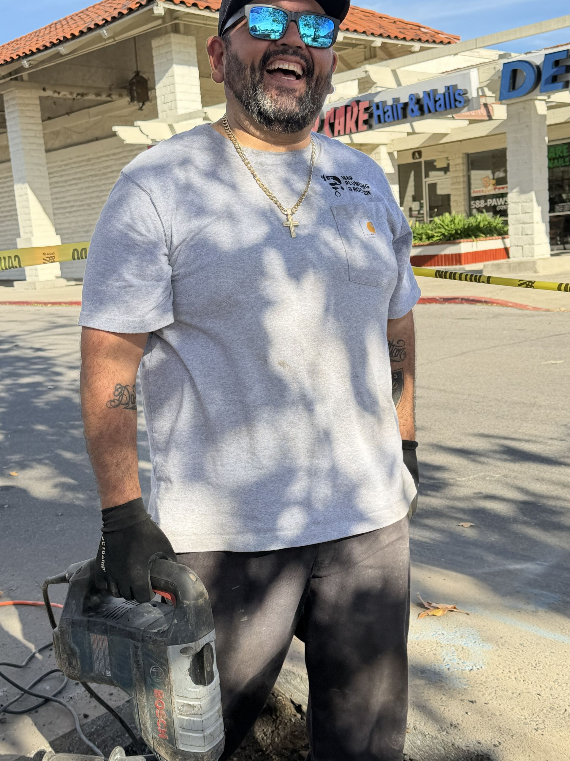 Man holding a power drill, wearing sunglasses and gloves, standing outside a storefront in sunlight.
