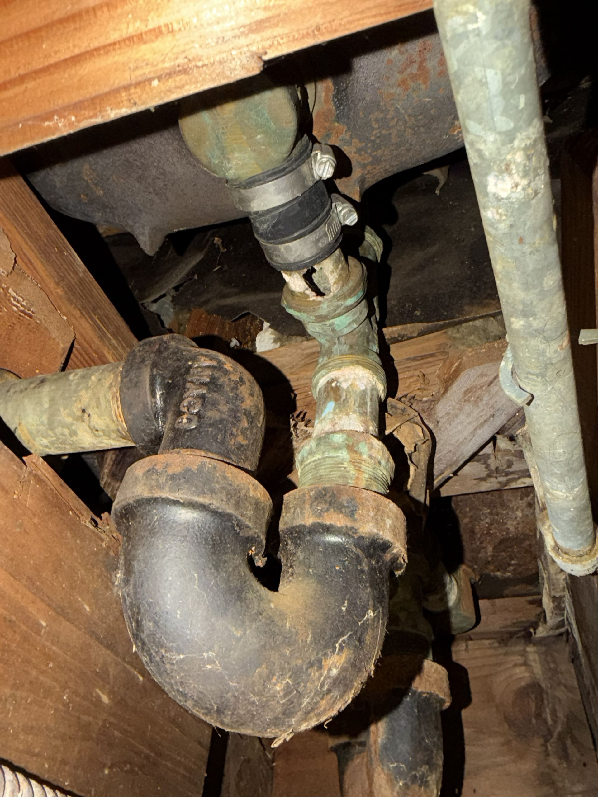 Old corroded metal plumbing pipes and joints under a wooden floor.