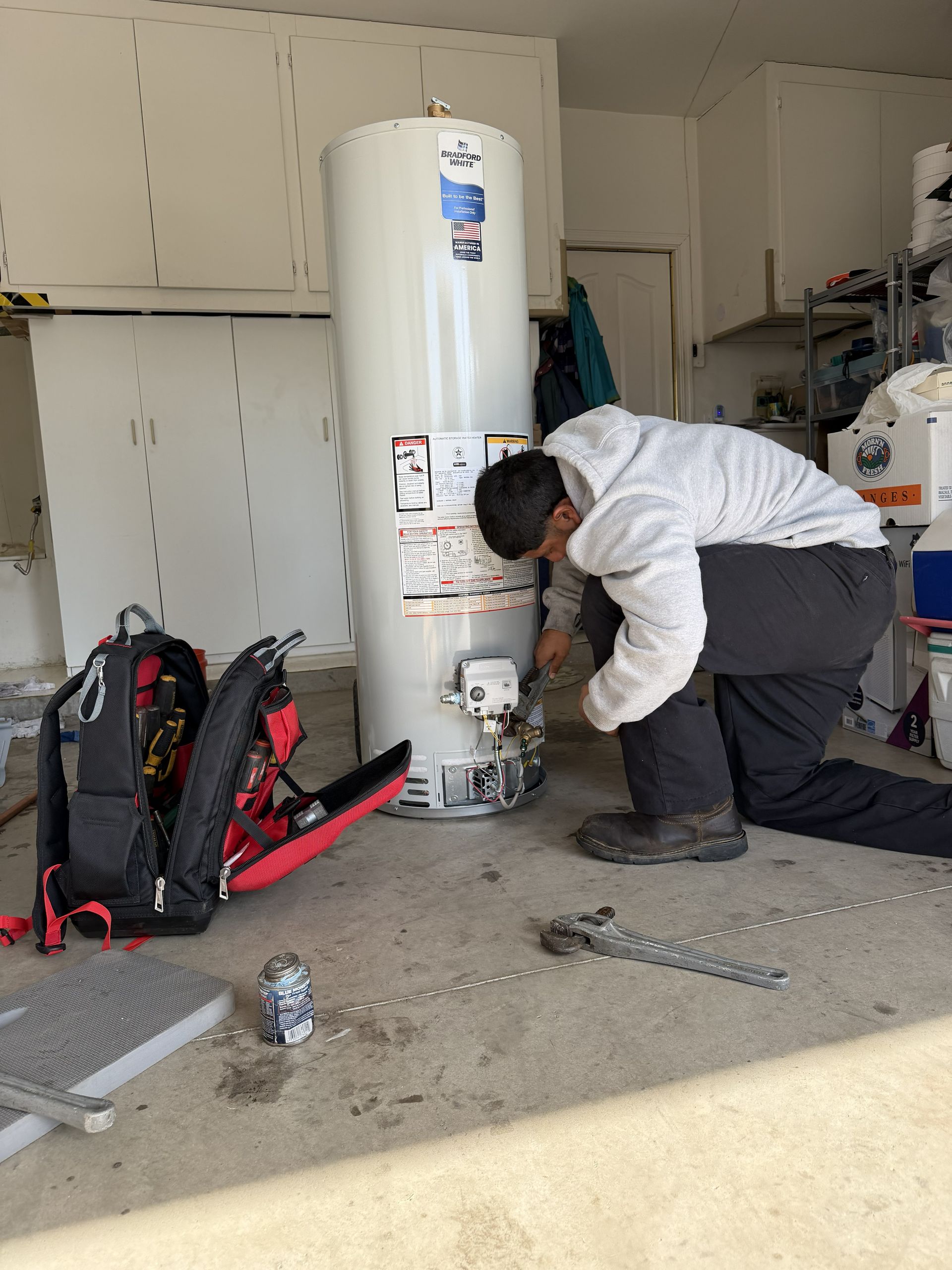 Technician kneels beside a water heater, inspecting pipes in a garage with tools and gear nearby.