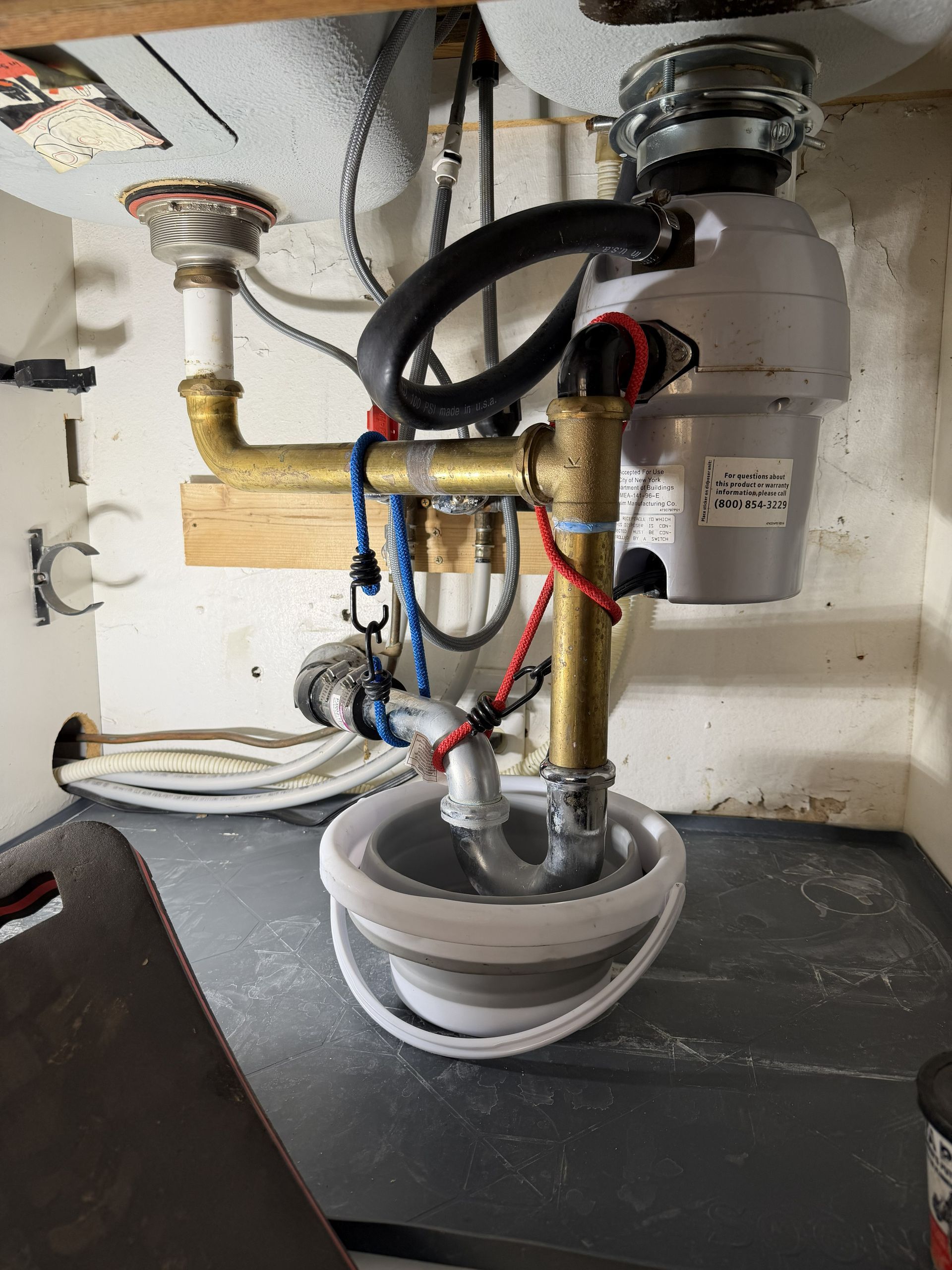 Under-sink plumbing with exposed pipes, valves, and a white bucket catching water or debris