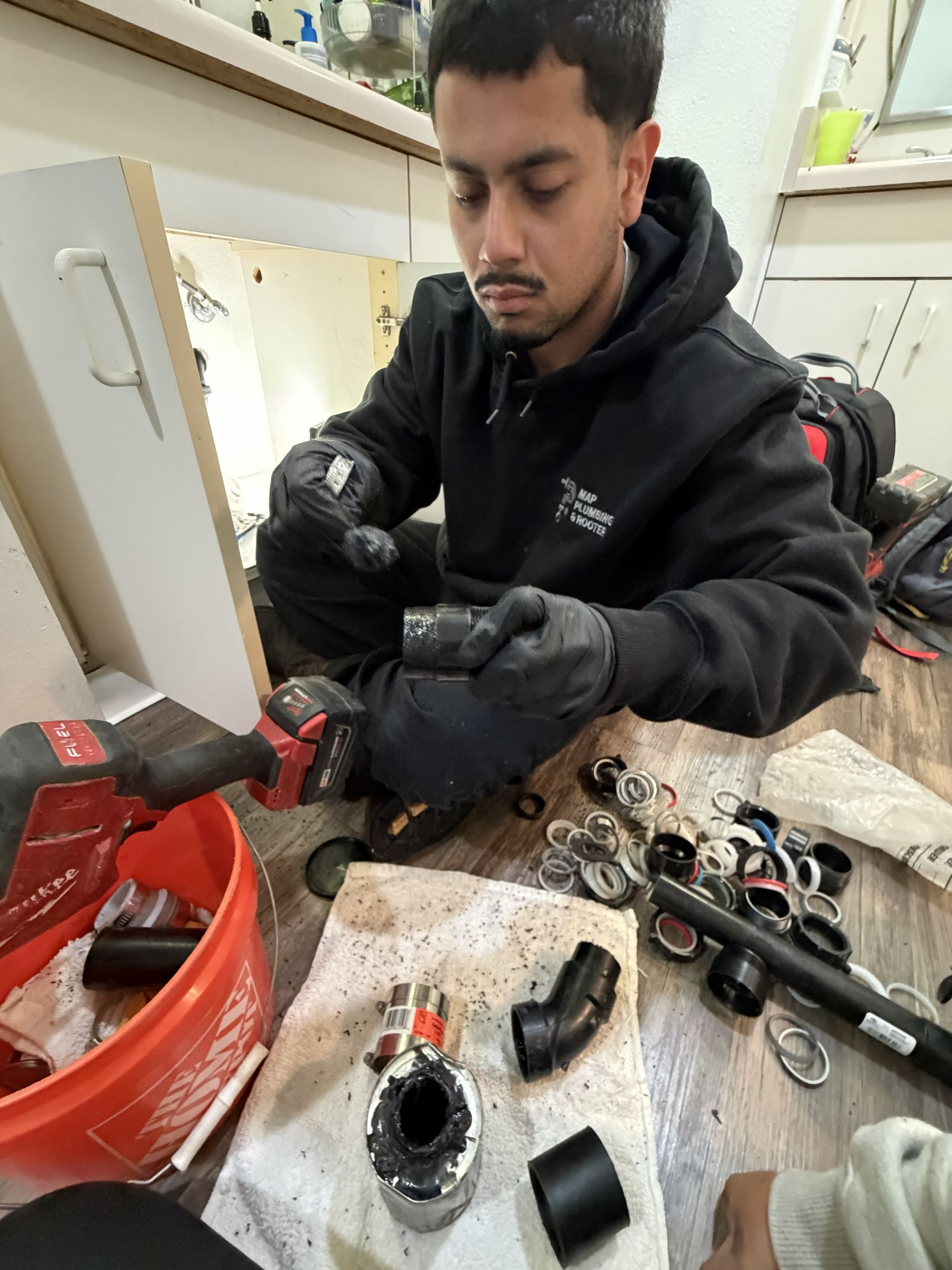 Mechanic in black hoodie assembling car parts at a workbench with tools and components scattered around.
