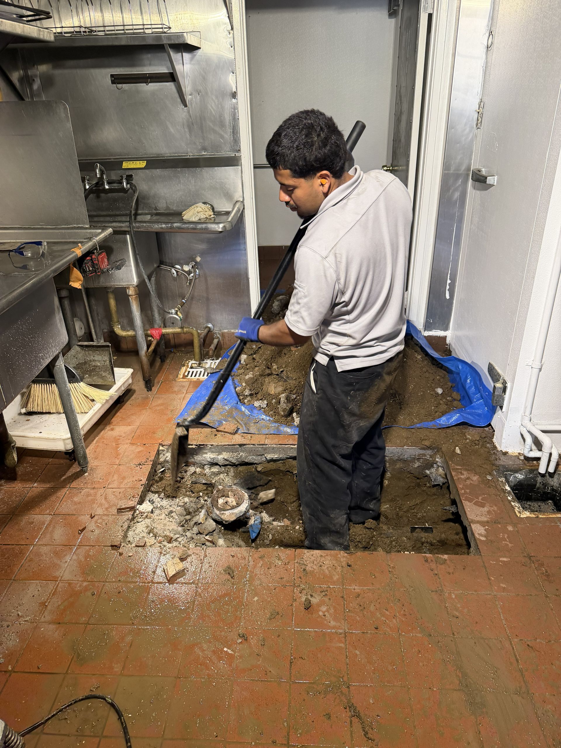 Worker shoveling debris in a wet industrial kitchen with blue tarp and shelves.