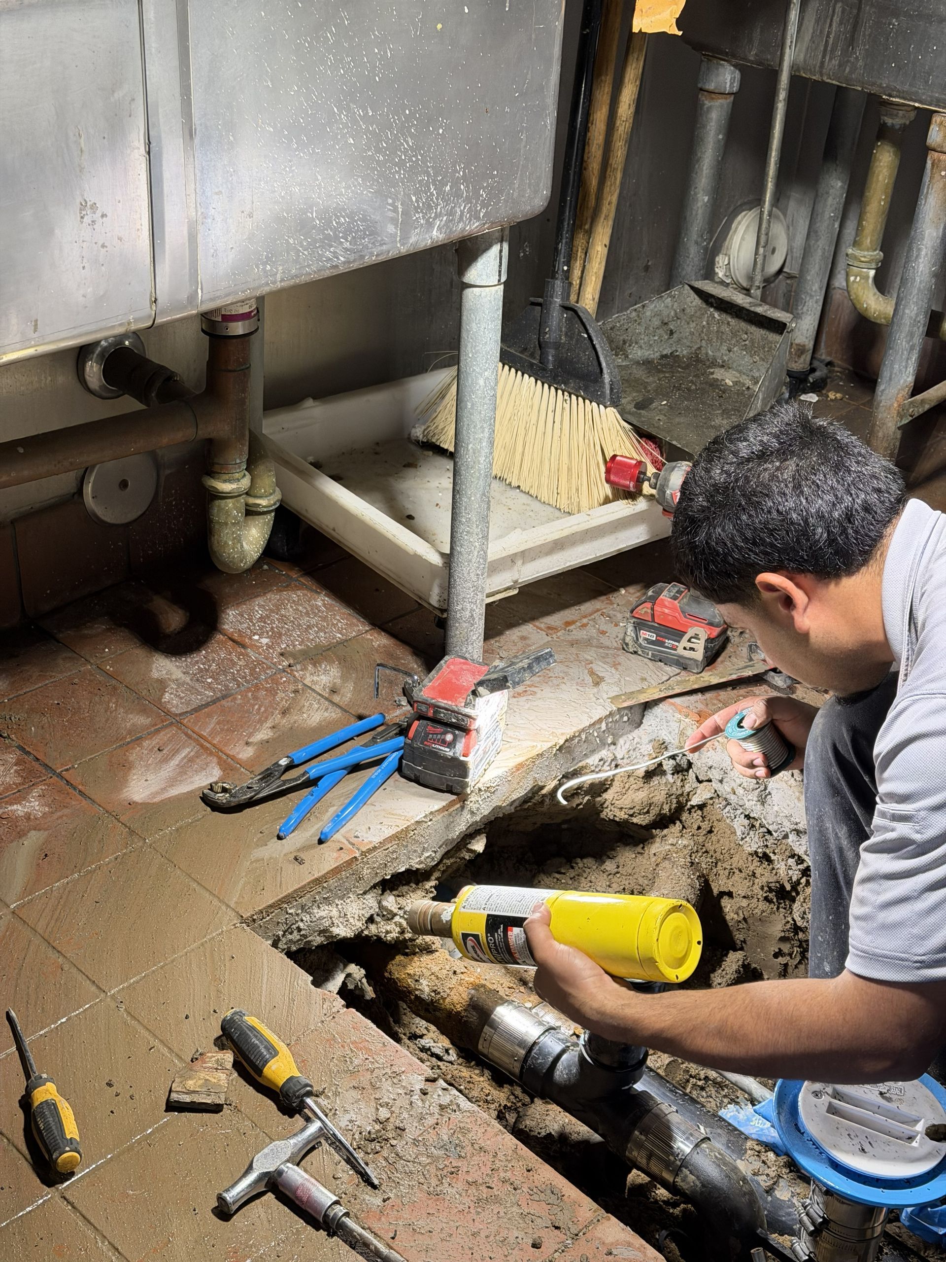 Worker repairing plumbing under a sink with tools on the floor and a yellow flashlight nearby