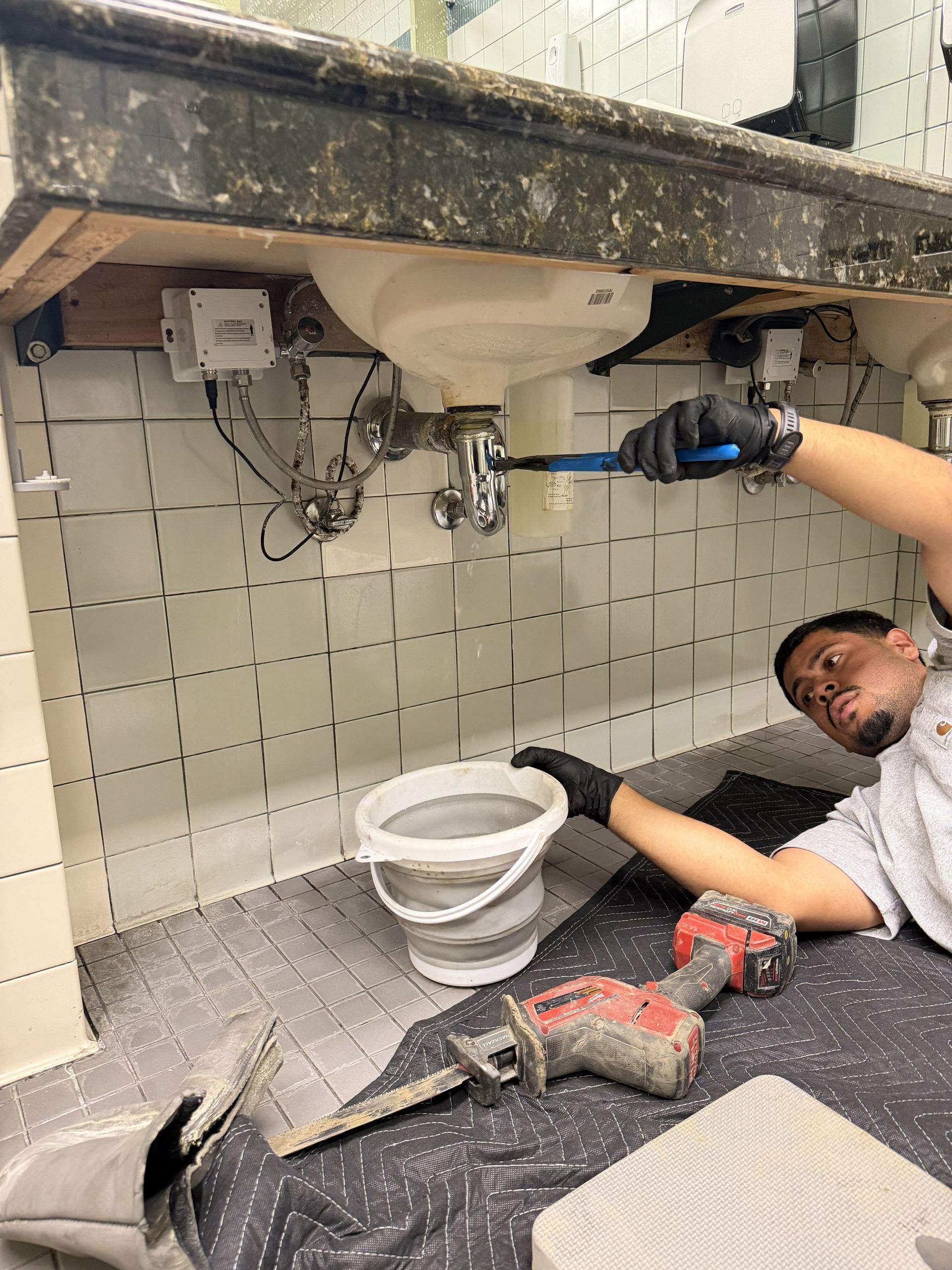 Person repairing plumbing under a sink, with tools and a bucket on the floor