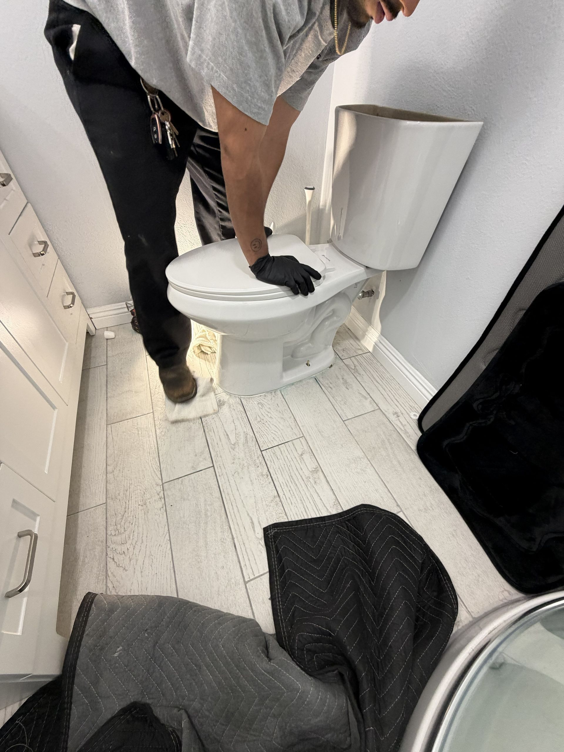 Person cleaning a white toilet in a narrow bathroom with gray walls and floor towel.