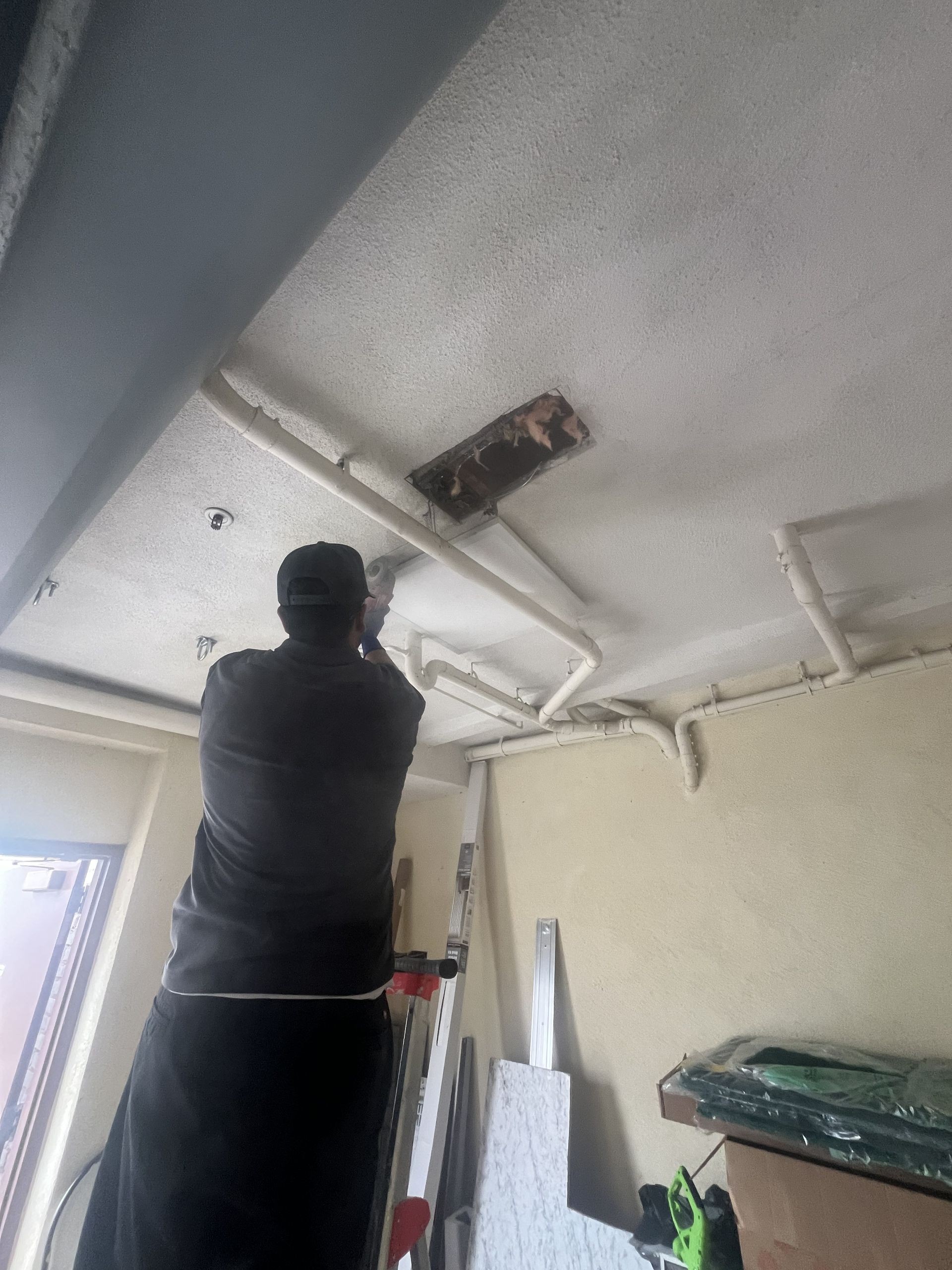 Person standing on a ladder, inspecting a damaged hole in a white ceiling inside a room.