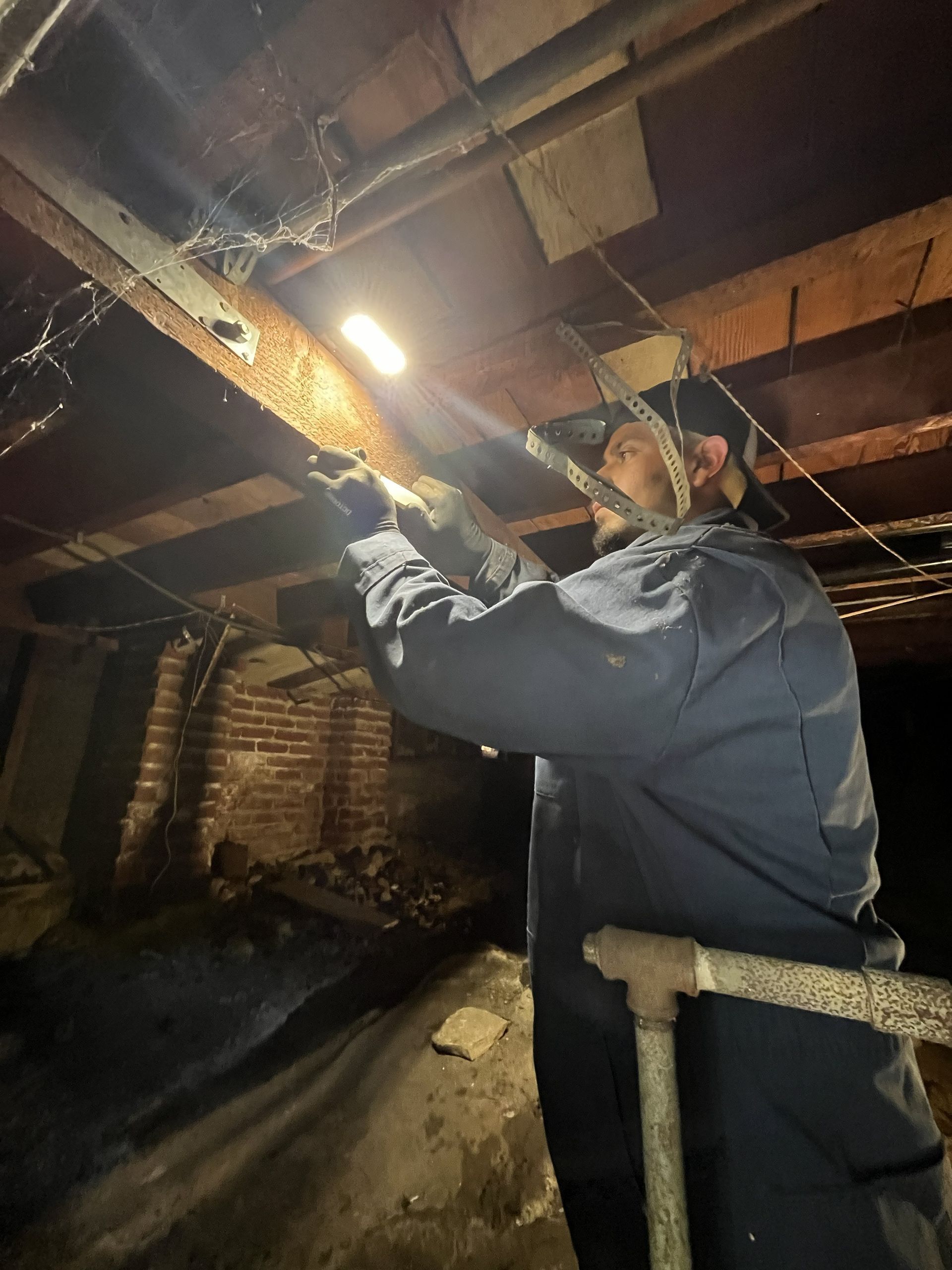 Worker inspecting exposed wooden beams in a dim basement with a bright flashlight.
