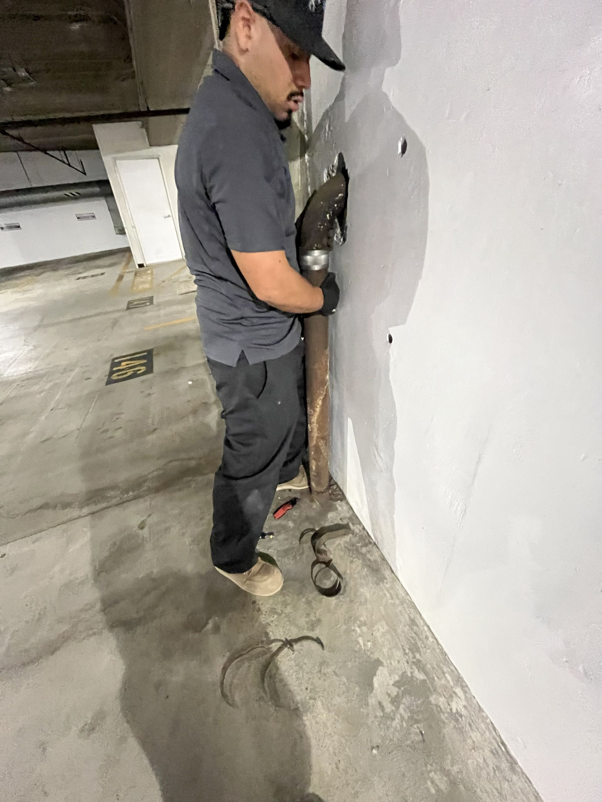 Worker using a jackhammer or drill along a concrete wall in a parking garage with debris on the floor