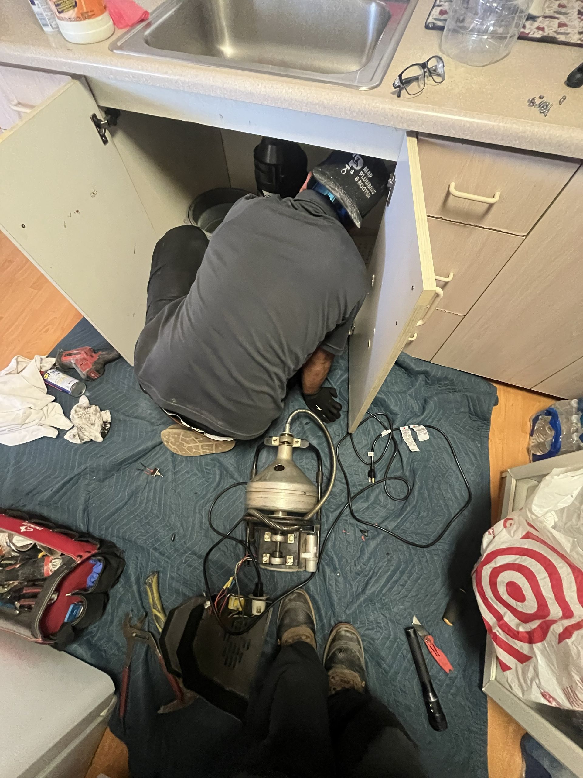 Person kneeling under a kitchen sink, working on plumbing amid tools and exposed pipes.
