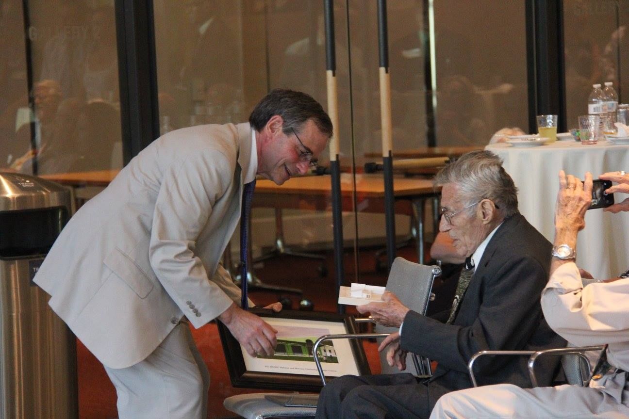Man in a beige suit leaning over a seated man in a dark suit at a table in a formal indoor setting